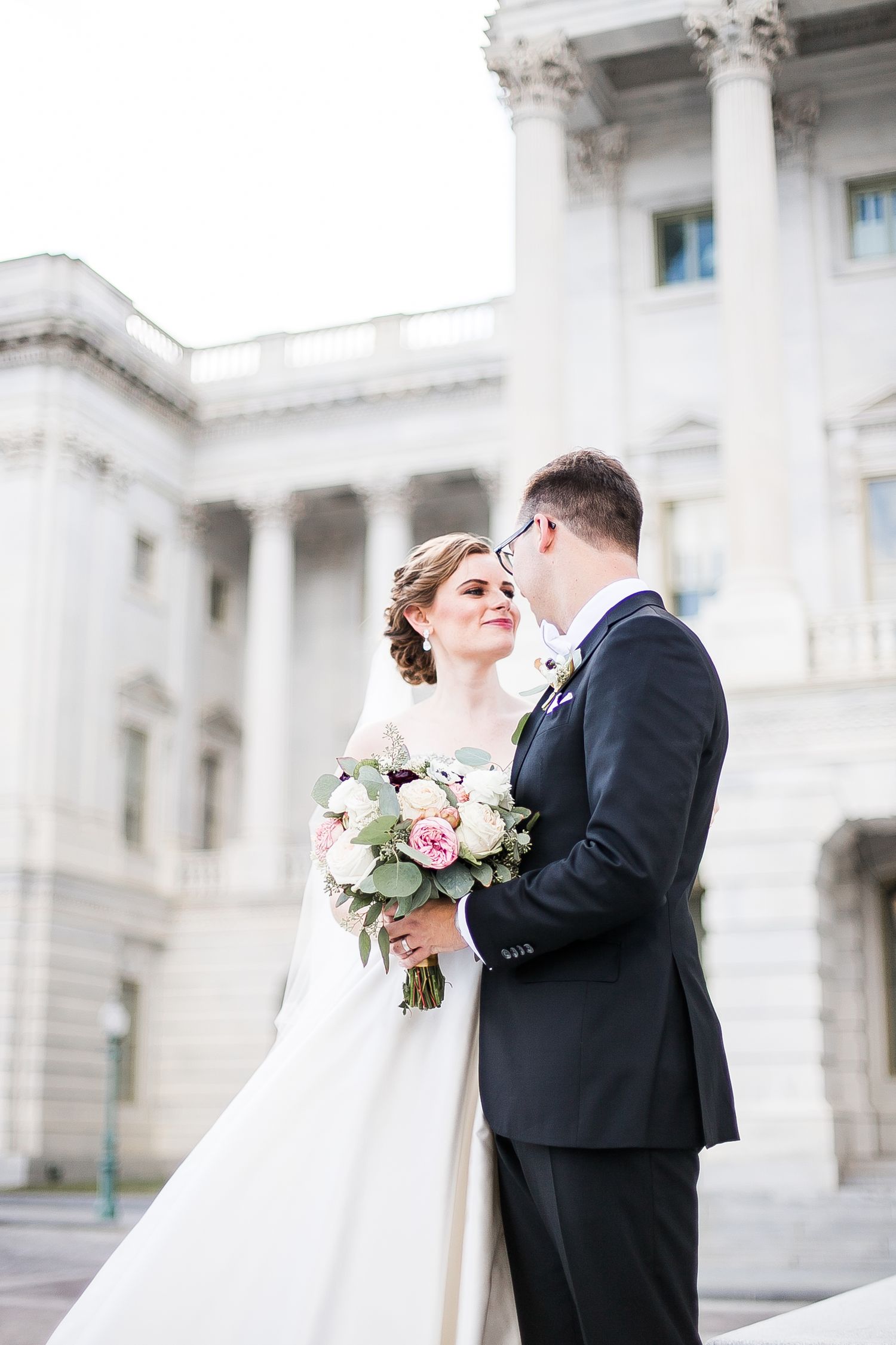 Romantic wedding couple shares an intimate moment in front of grand white stone architecture.