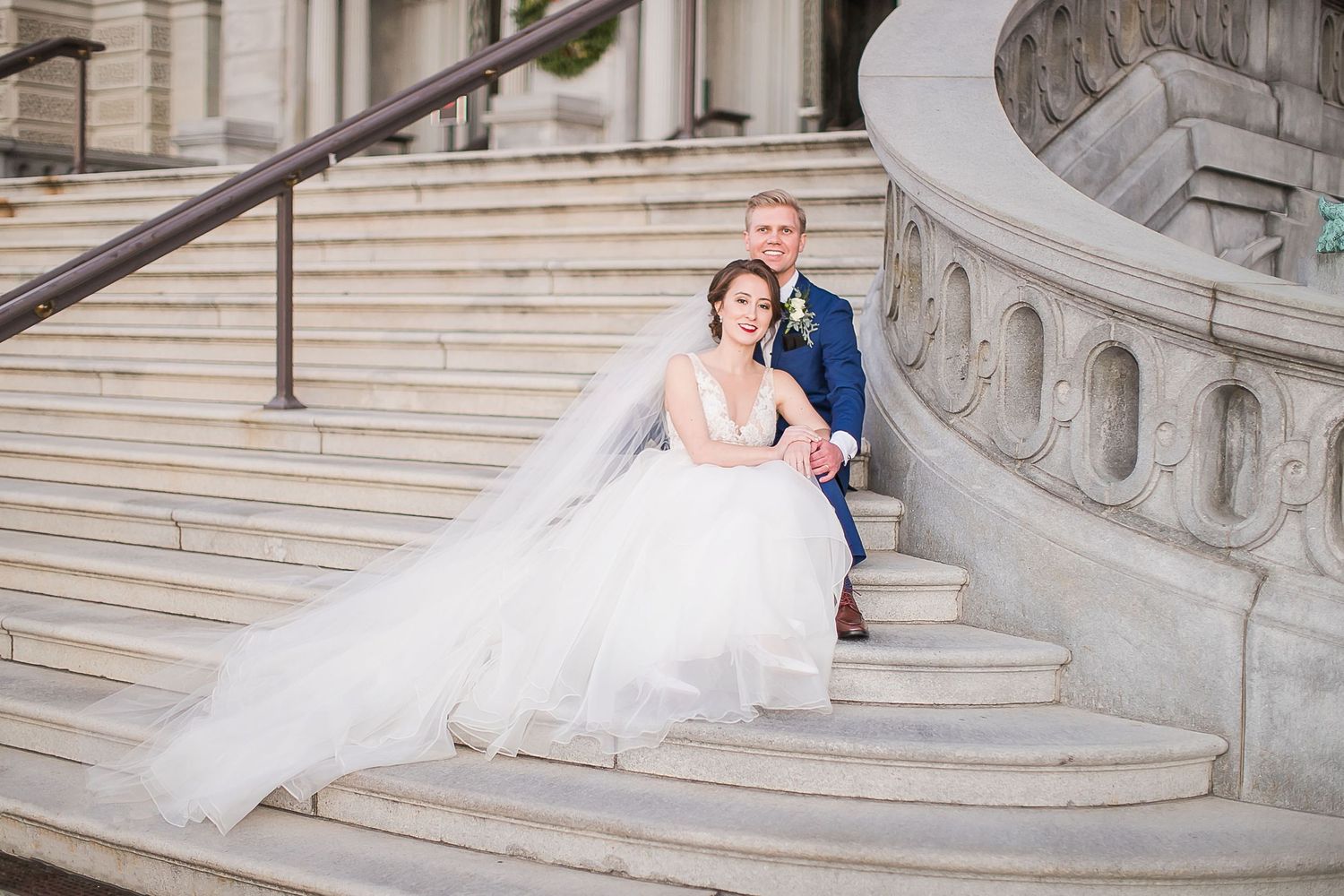 A bride in a flowing white gown and groom pose together on elegant stone steps of a grand building.