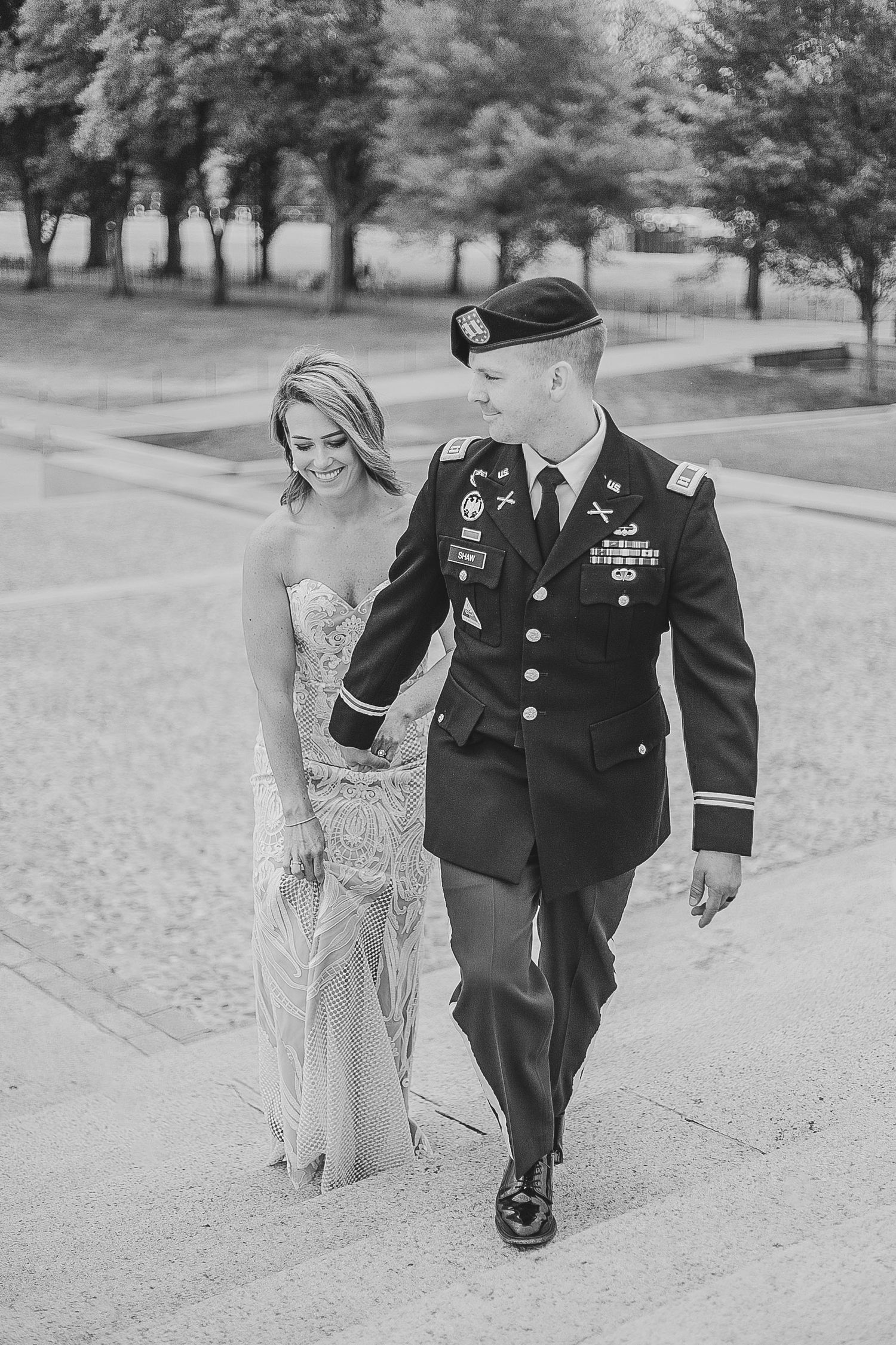 A couple walking hand in hand on a path, with one in a formal military dress uniform and the other in an elegant gown.