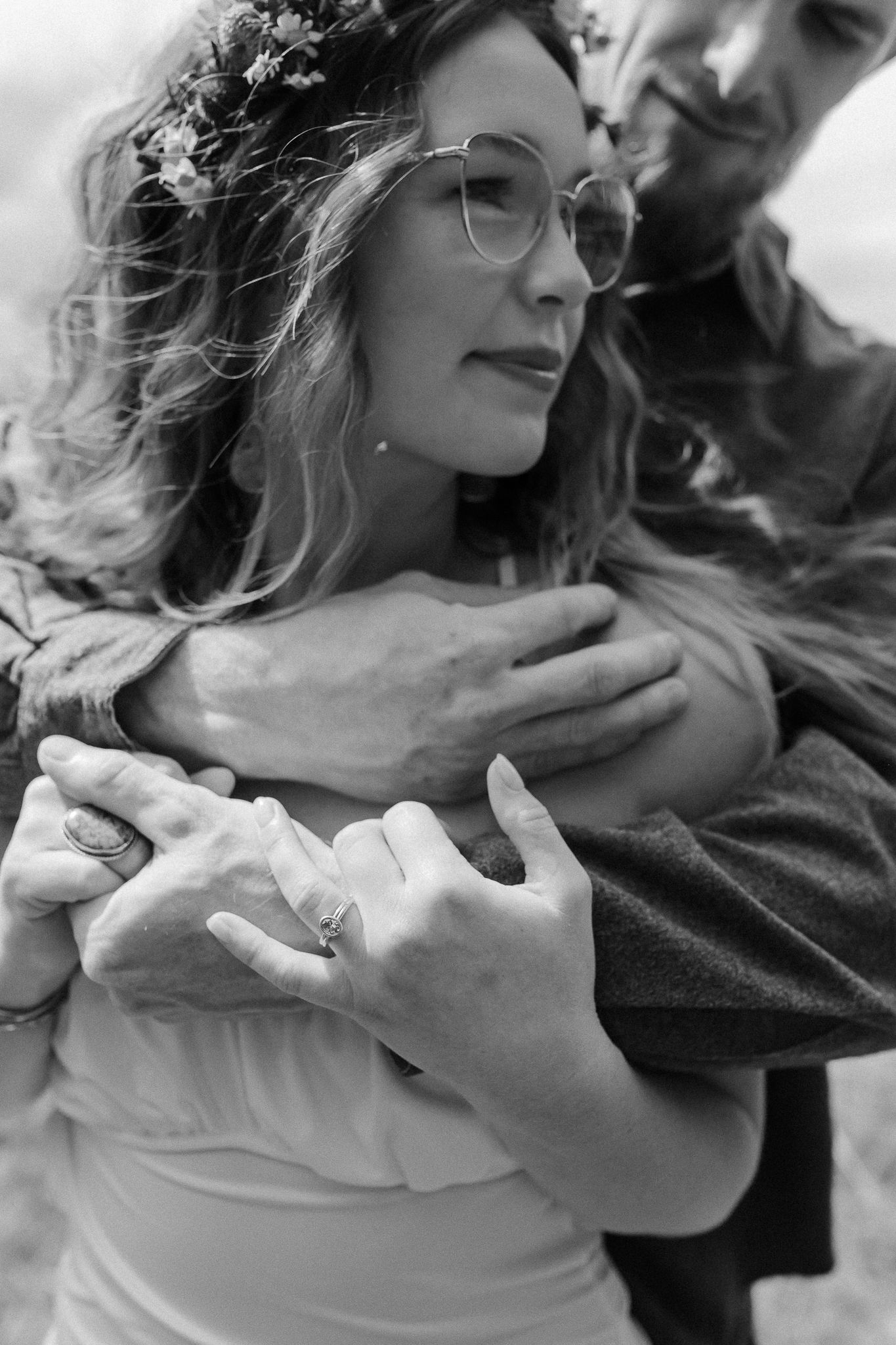 Intimate black and white portrait of a couple embracing, with focus on round glasses and flowing hair in natural light.