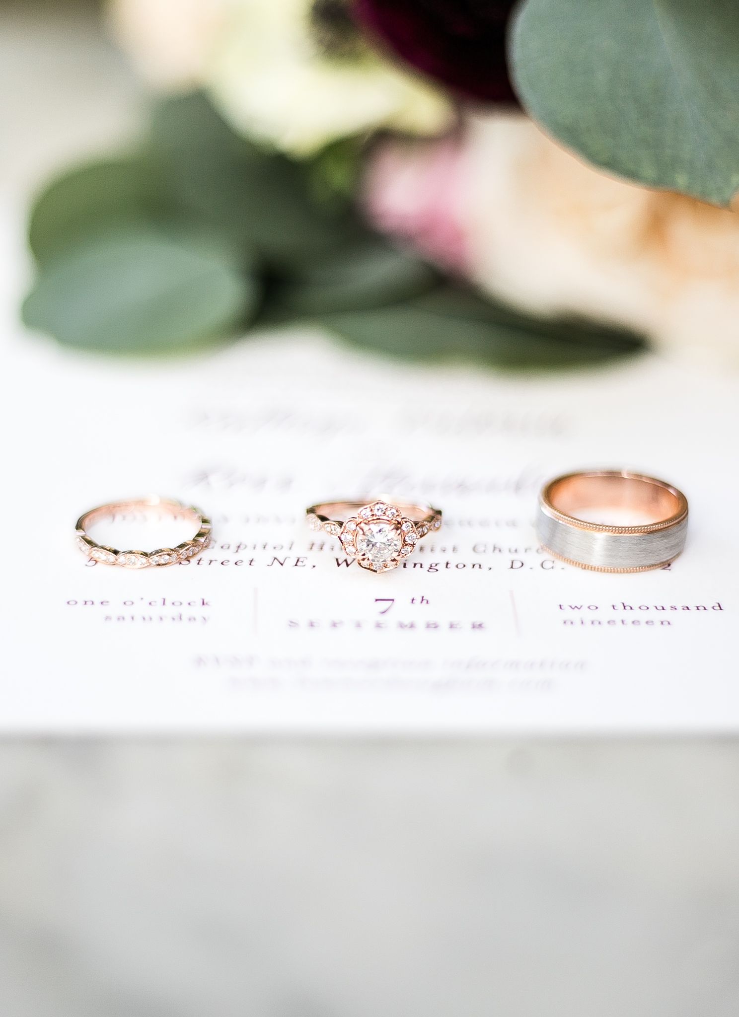 Three delicate rose gold wedding rings with intricate details displayed on a white surface with green foliage accents.
