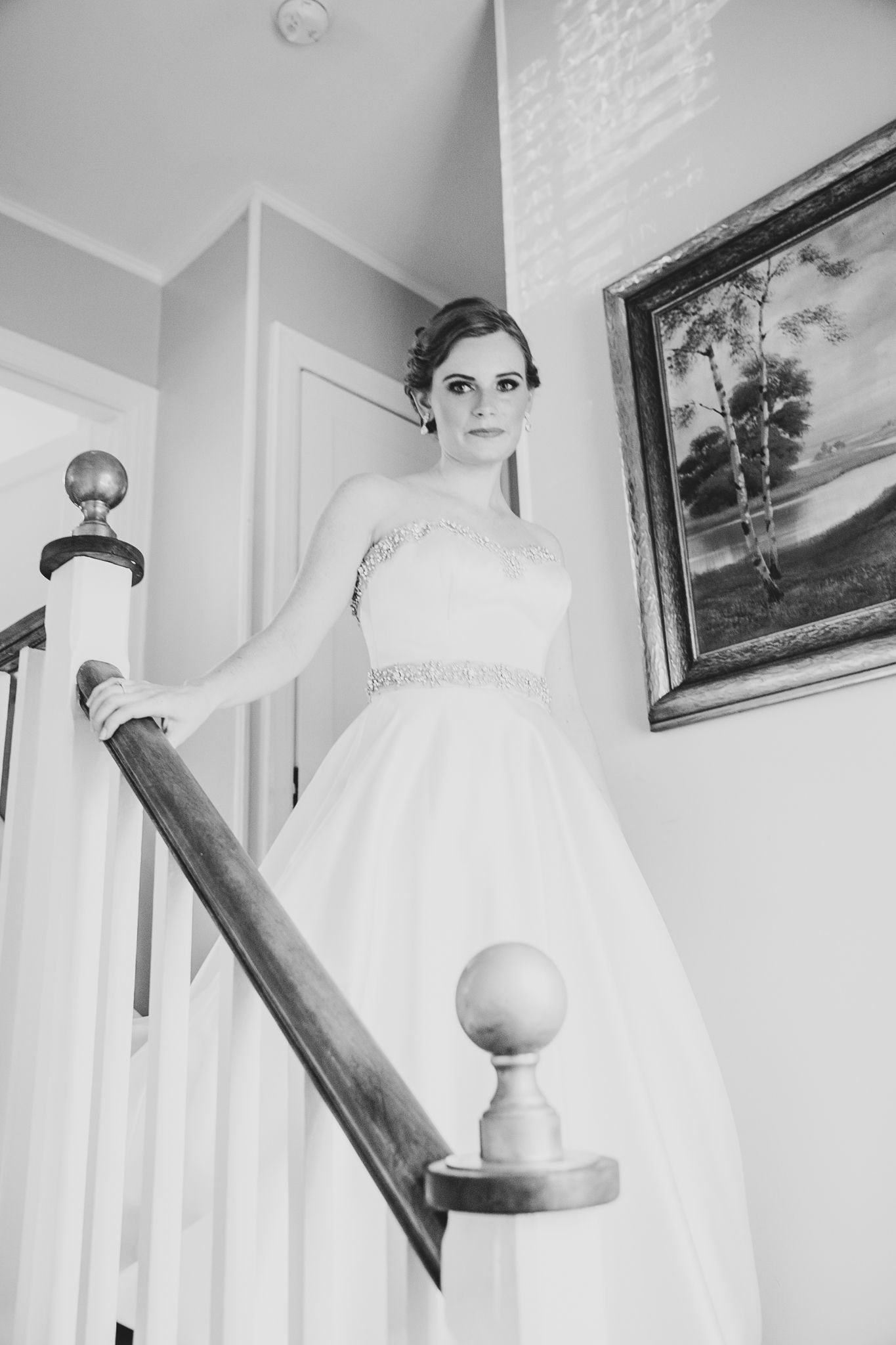Black and white photo of a bride in an elegant white wedding gown with beaded detail, standing on stairs by a bannister.