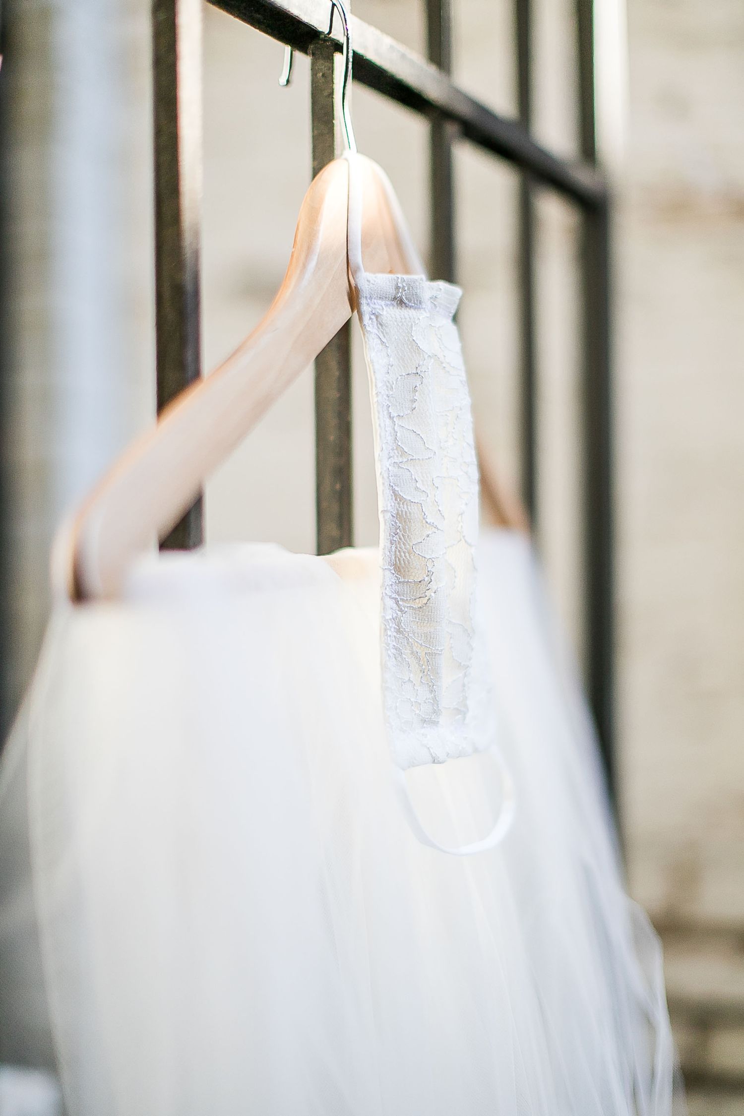 A delicate white wedding dress hangs on a blush pink hanger against a soft blurred background.