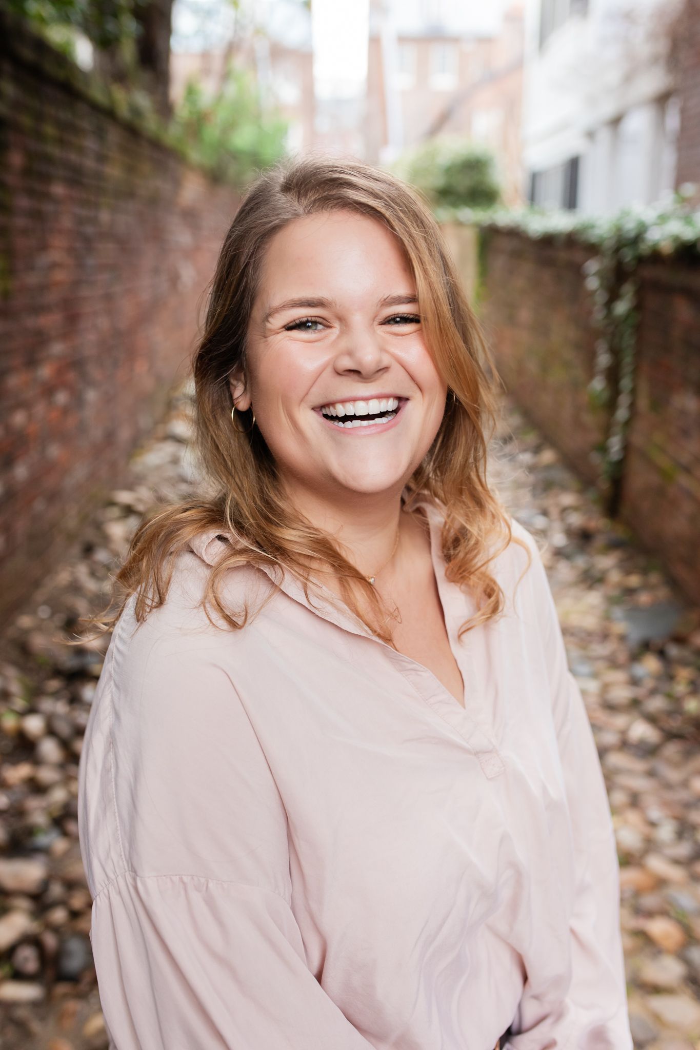 A person wearing a light pink button-up shirt smiles warmly while posing for a portrait photo shoot against a brick wall.