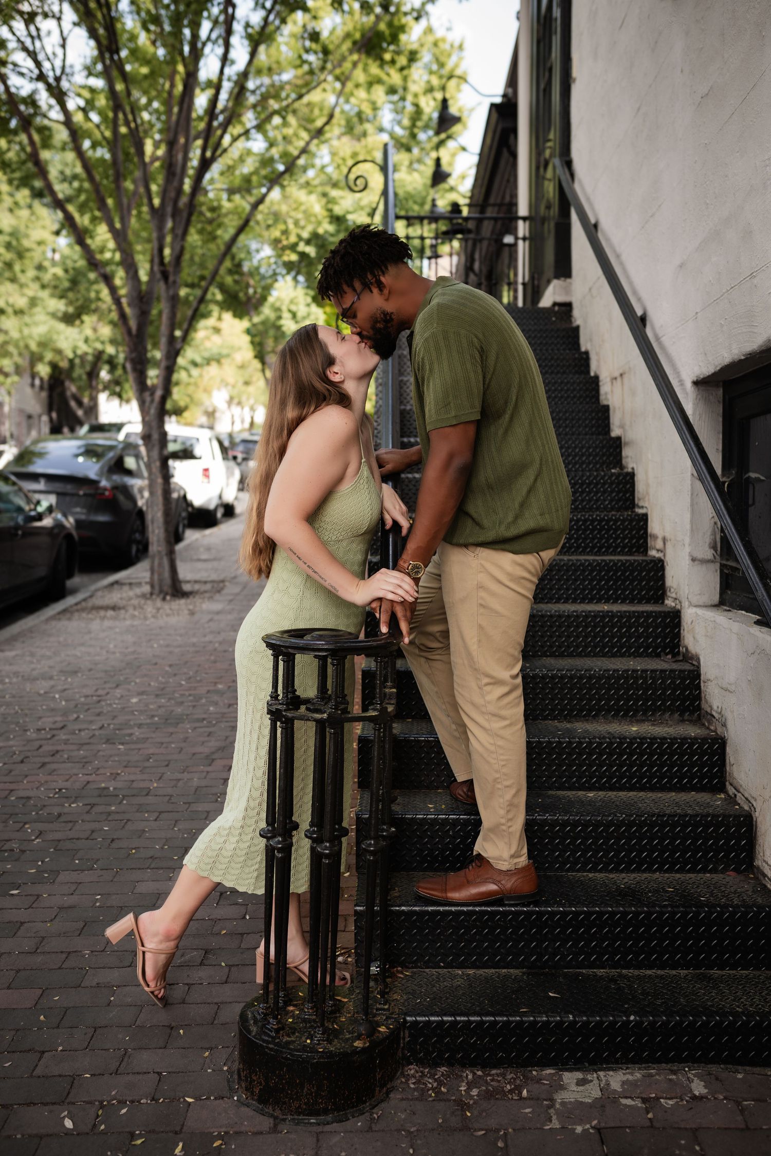 A romantic couple share a tender moment on an outdoor staircase in front of a brick sidewalk lined with trees.