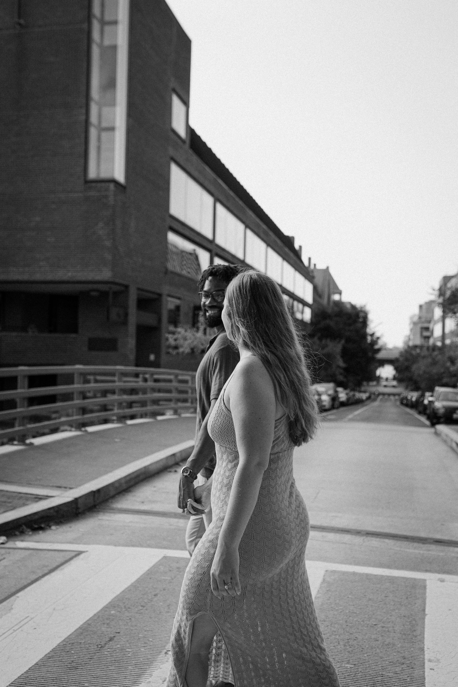 Black and white street photograph of two people walking across a crosswalk in an urban setting at dusk.