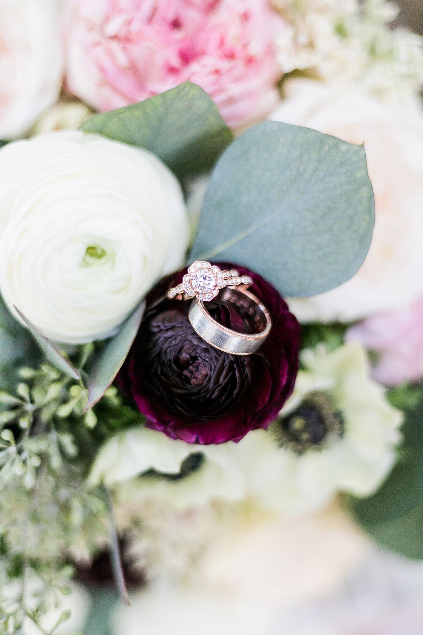 Delicate engagement ring with diamonds rests on a deep purple ranunculus bloom amid a white and pink floral bouquet.