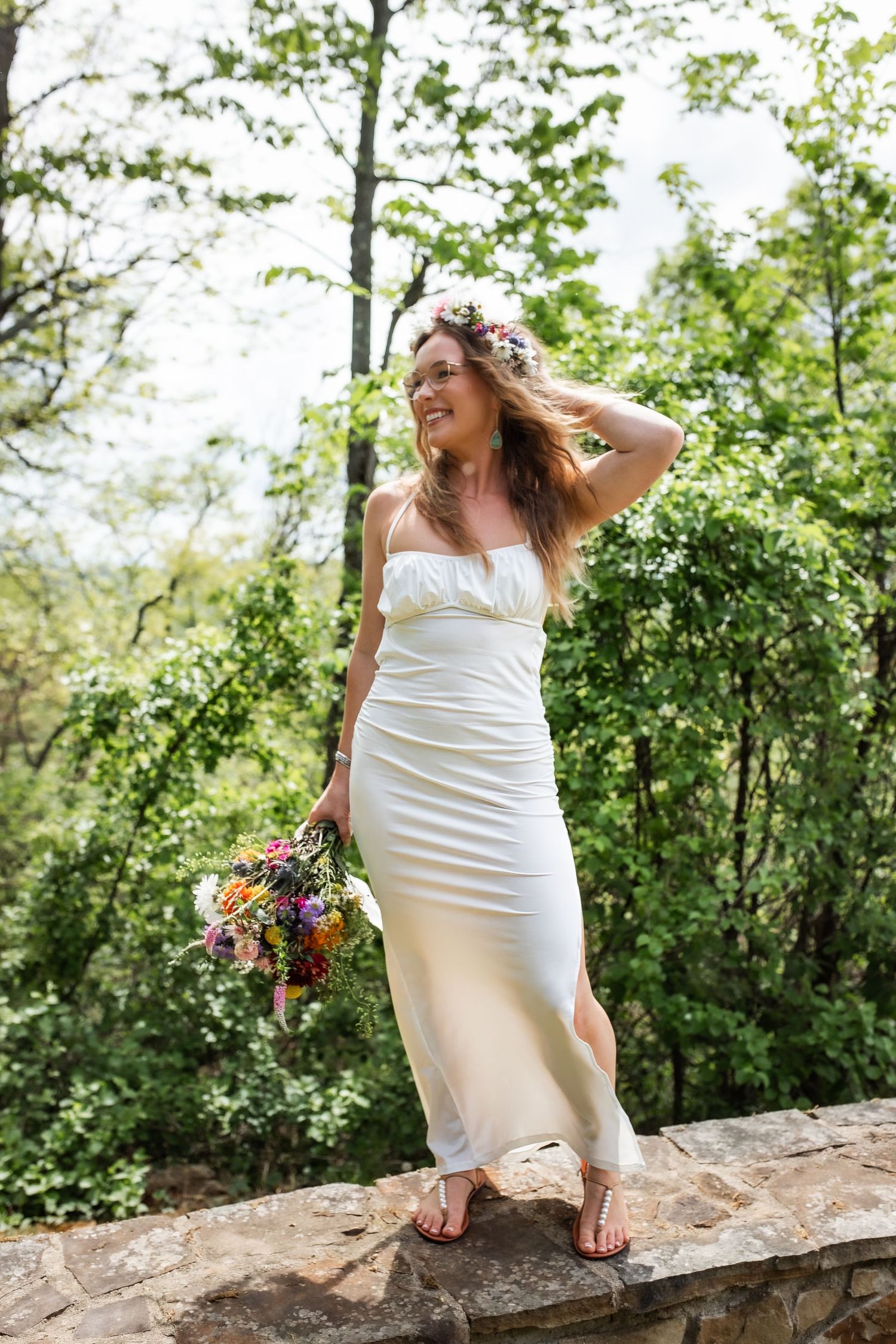Someone in a white wedding dress stands on a stone wall holding a colorful bouquet in a lush outdoor setting.