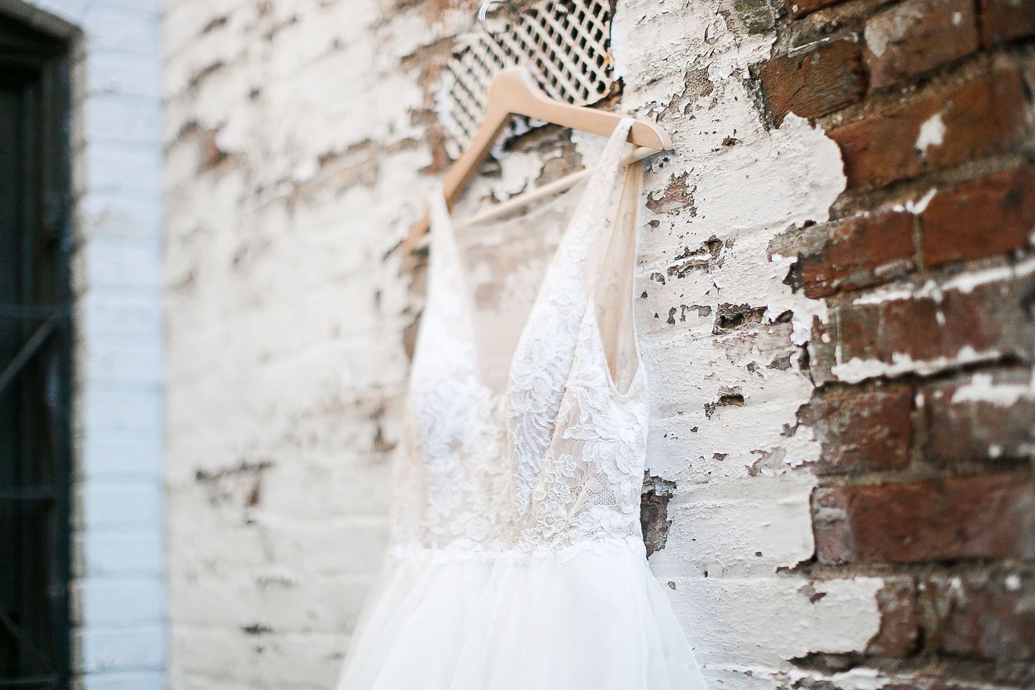 A delicate white lace wedding dress hangs against a distressed brick wall with peeling paint.