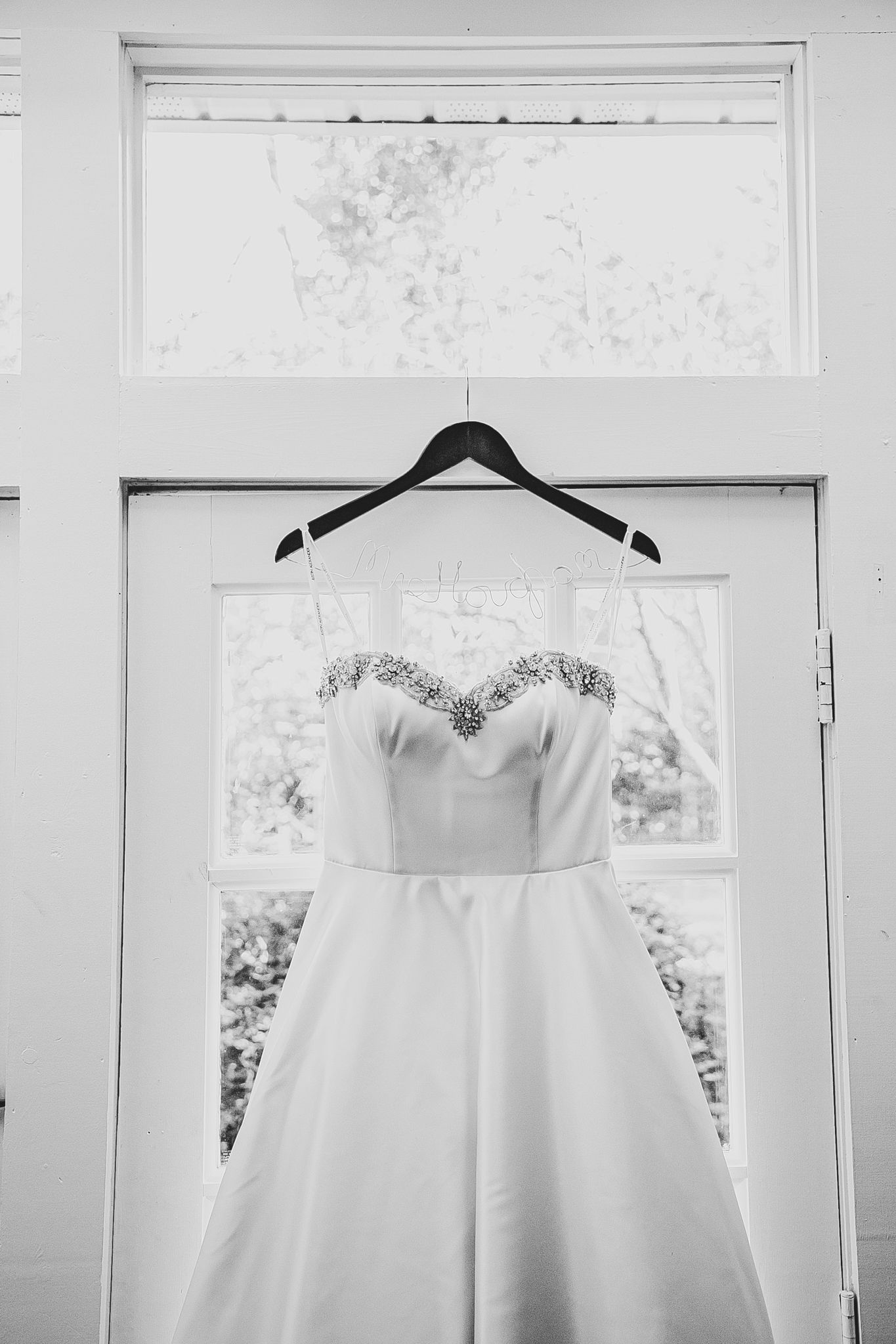 A black and white photograph showing an elegant white wedding dress hanging from a clothes hanger in a window.