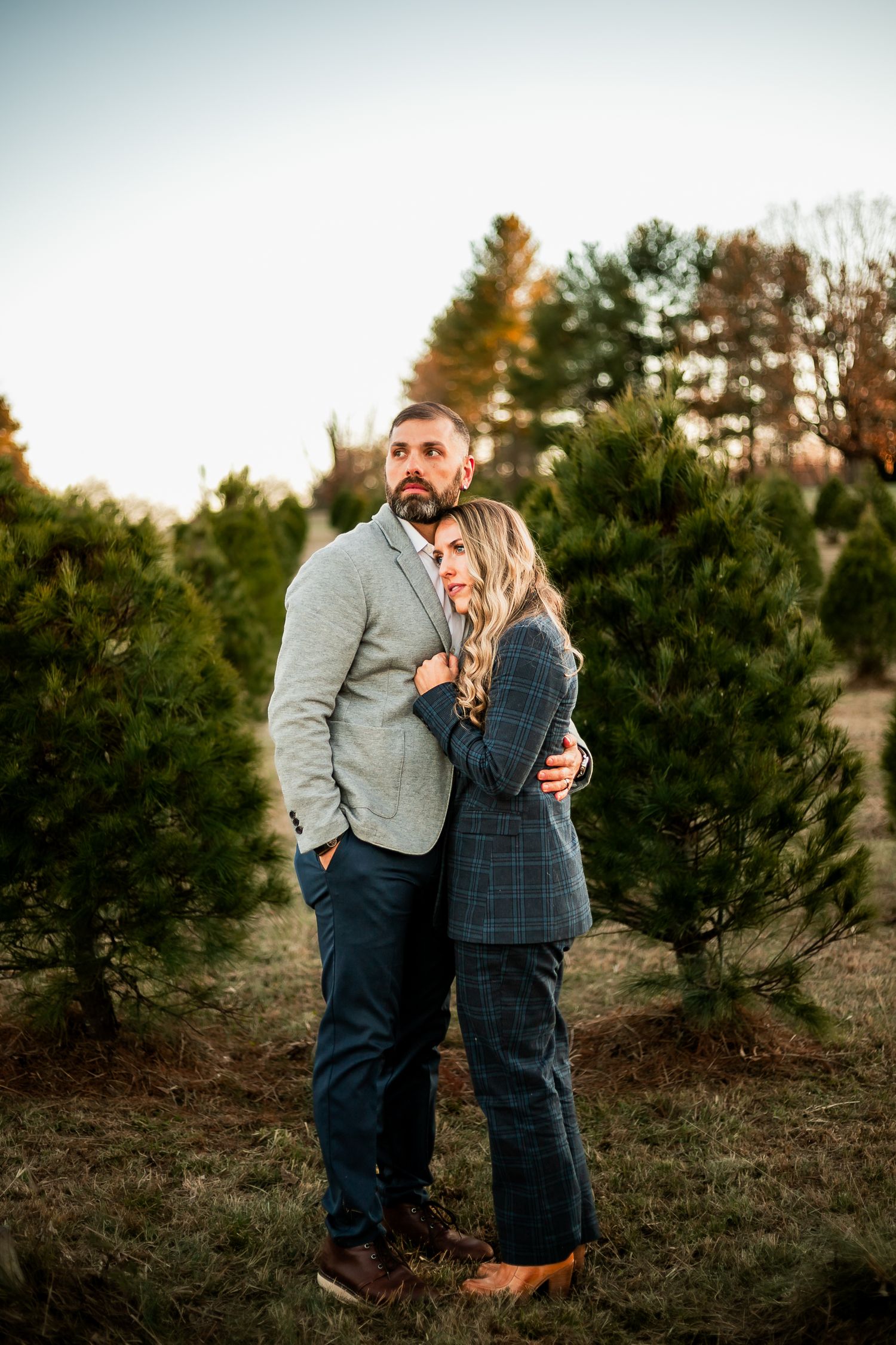 A couple shares a romantic moment among evergreen trees at a Christmas tree farm during sunset.