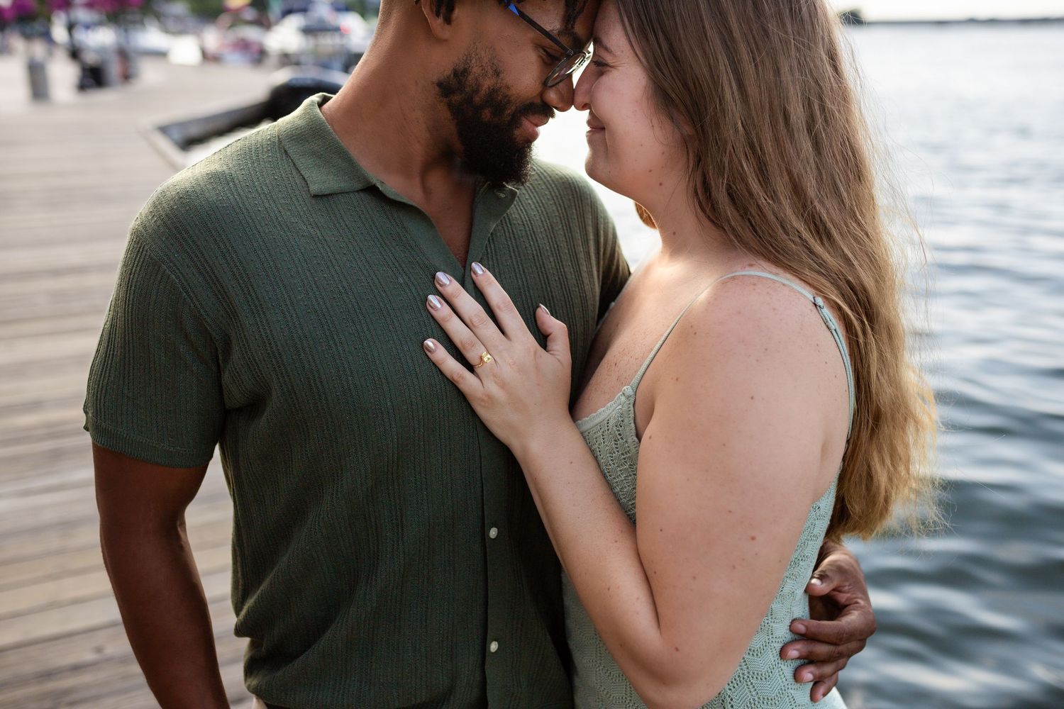 Romantic couple embraces by the waterfront as sun sets over marina in background.