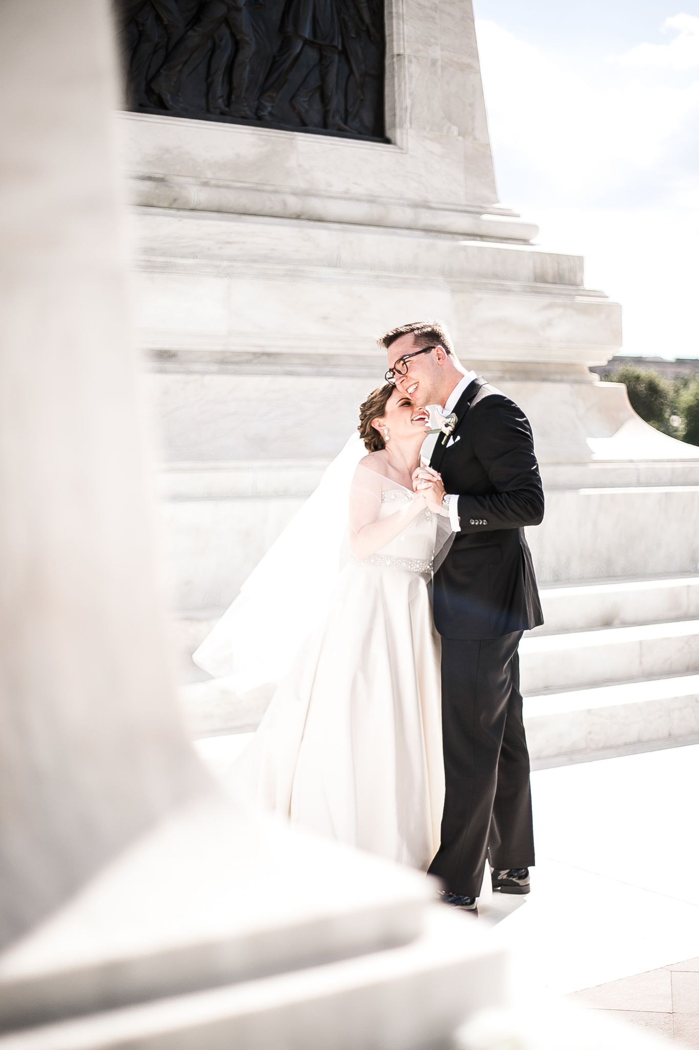A romantic wedding couple shares a tender moment on white marble steps in bright sunlight.