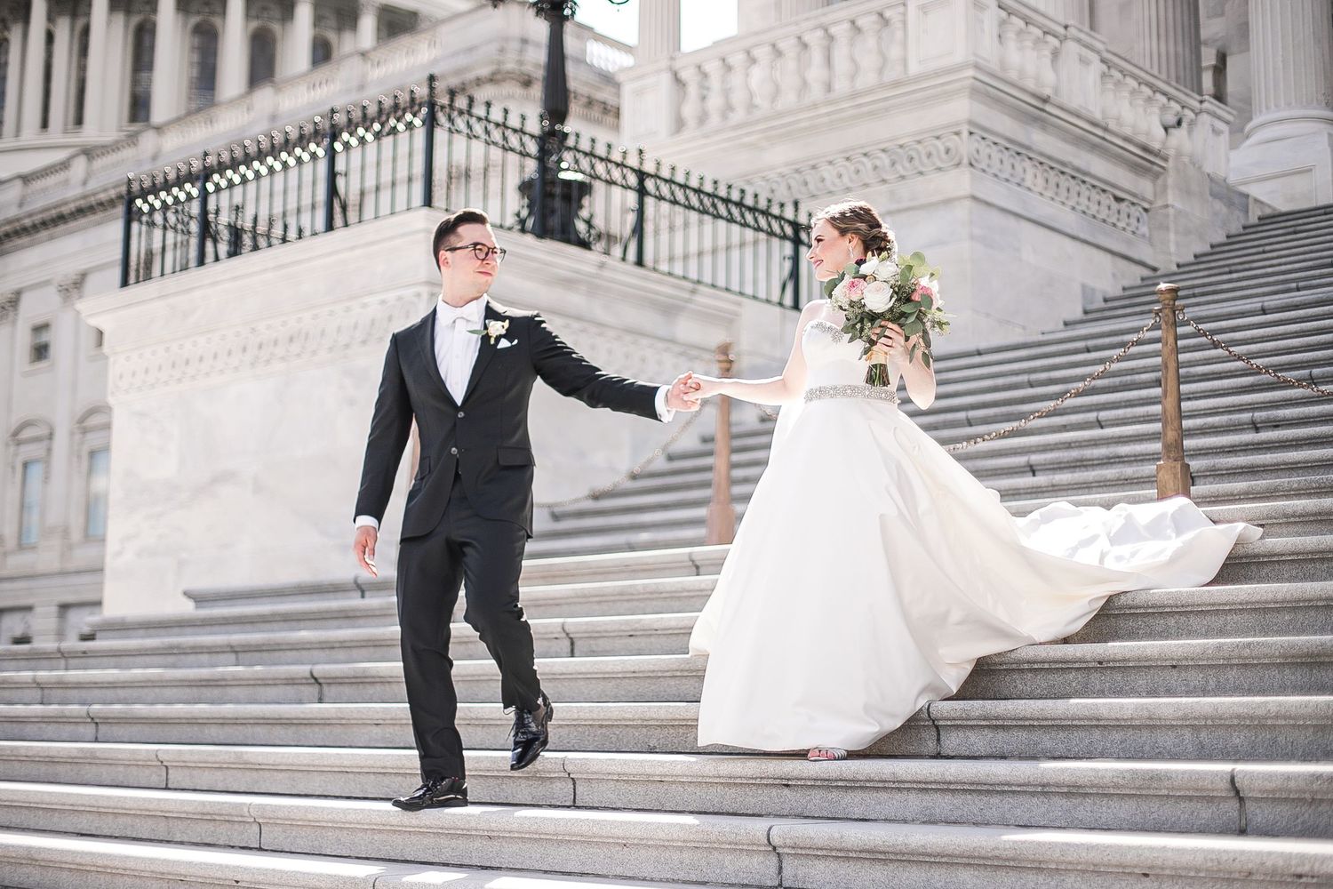 A couple in wedding attire performs playful dance moves on the grand marble steps of a classic architectural building.
