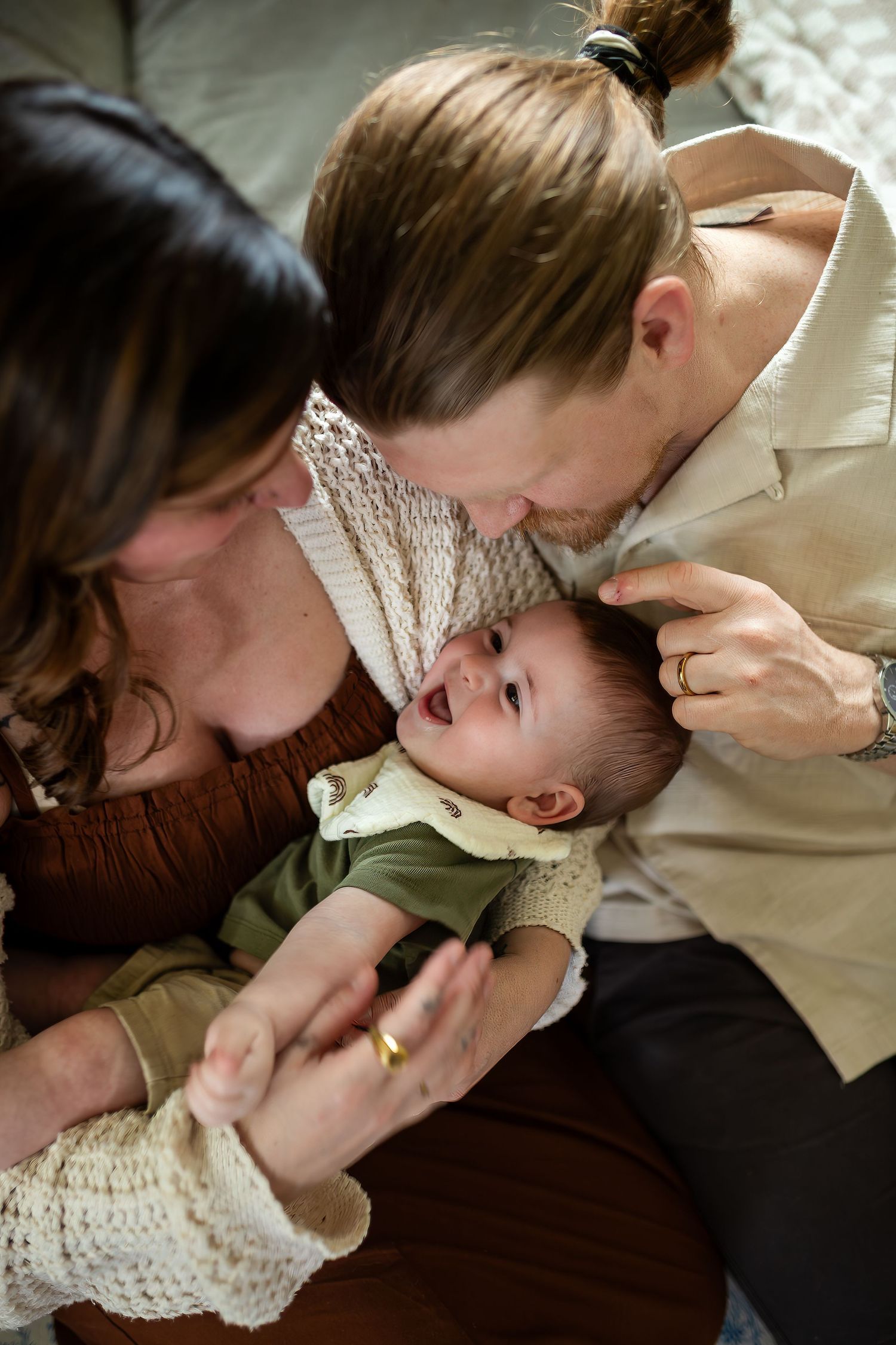 A parent kisses and shows affection to a small baby wearing a white knitted dress while lying on a dark surface.