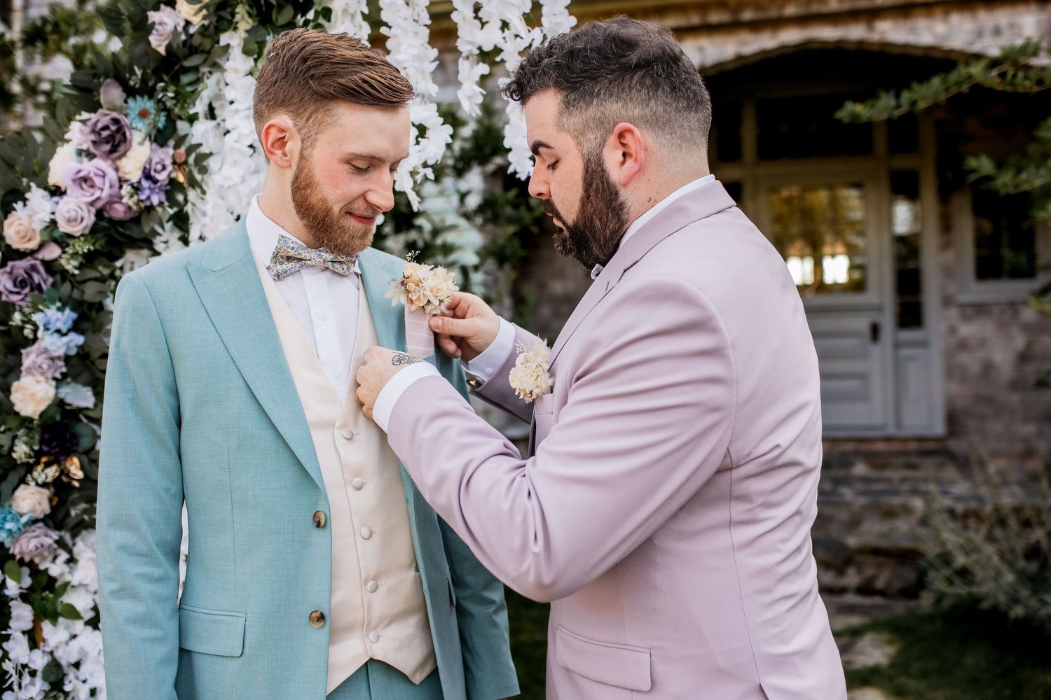 Two grooms in mint and pink suits adjusting boutonnieres under floral arch.