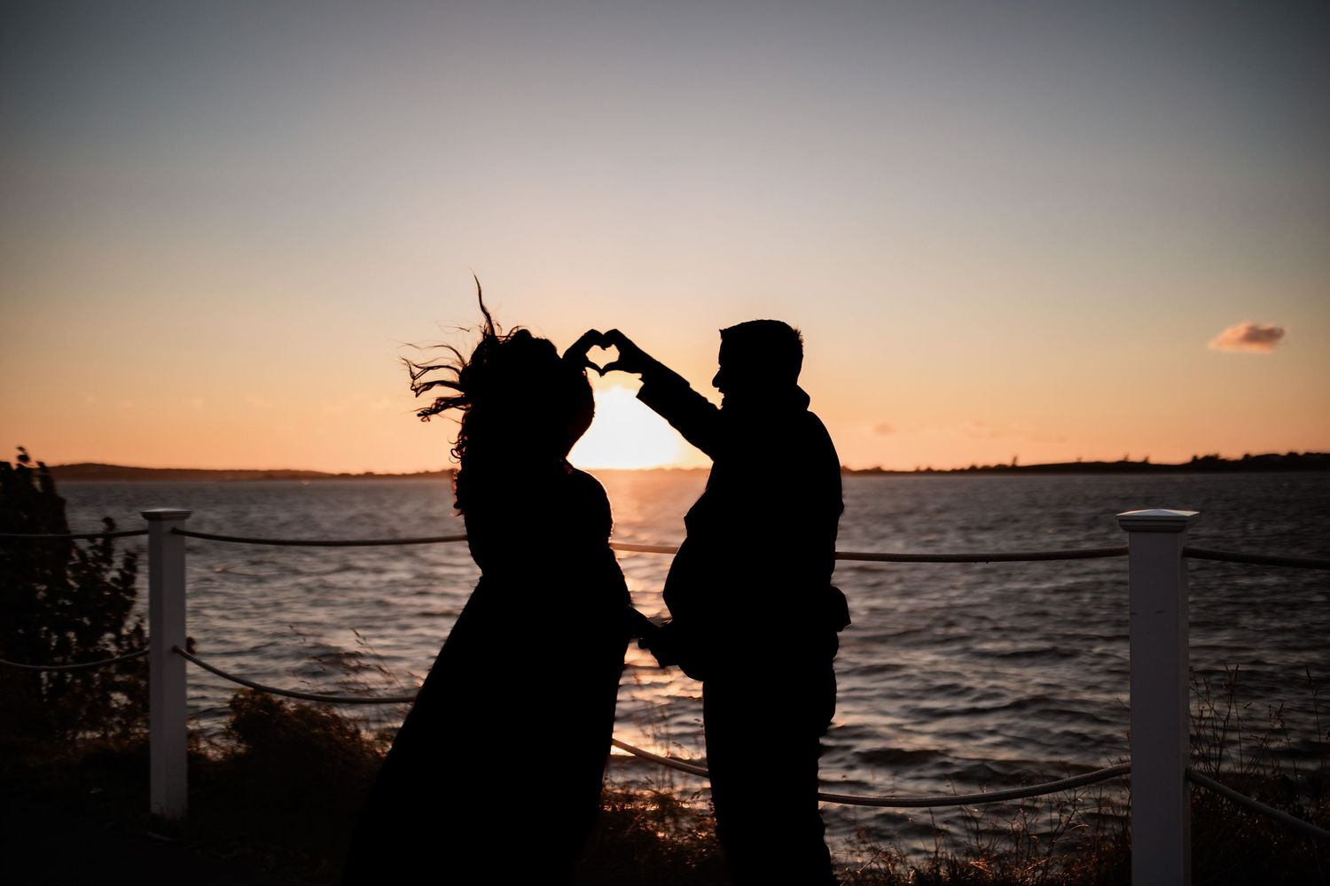 A romantic silhouette of a couple making heart shape with hands against sunset over water.