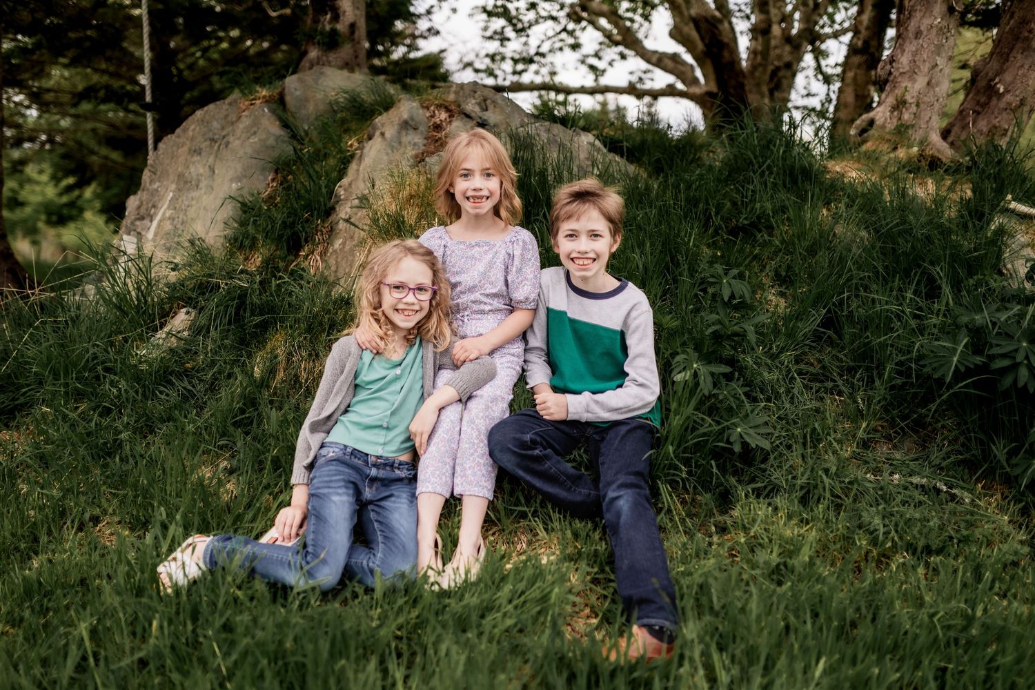Casual family portrait of group sitting together on grassy slope in natural outdoor setting.
