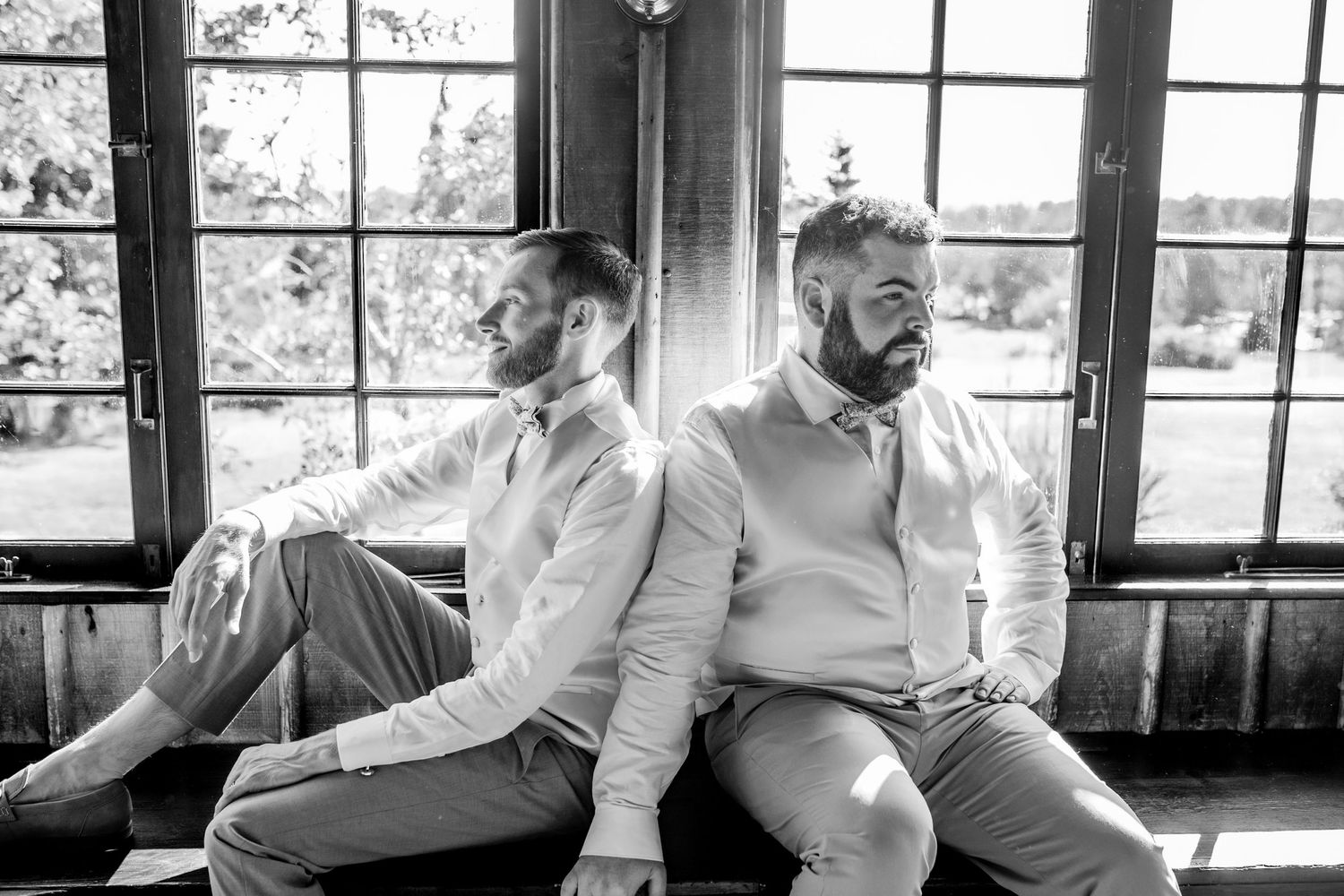 Black and white portrait of two men sitting back to back by window.