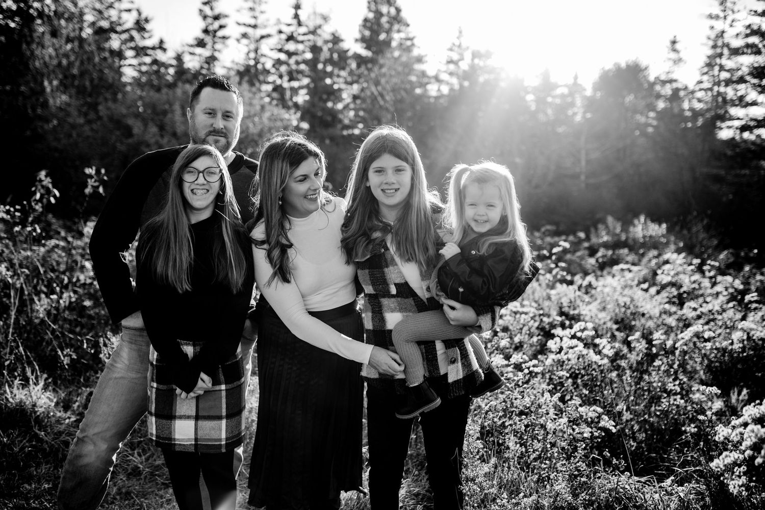 A black and white portrait of a group holding each other close while standing in front of evergreen trees.