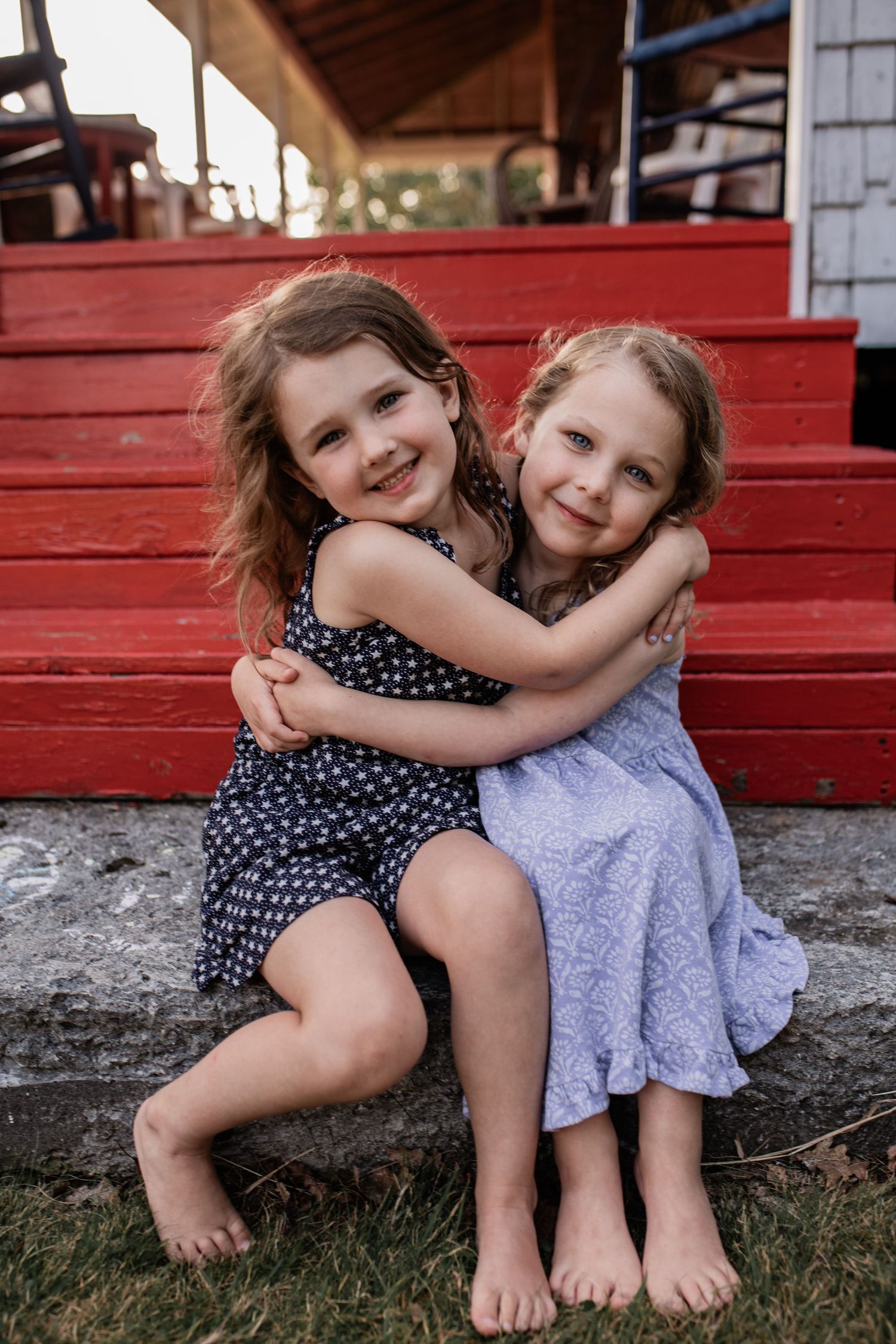 Two young children in summer dresses hugging and smiling on red painted wooden steps.