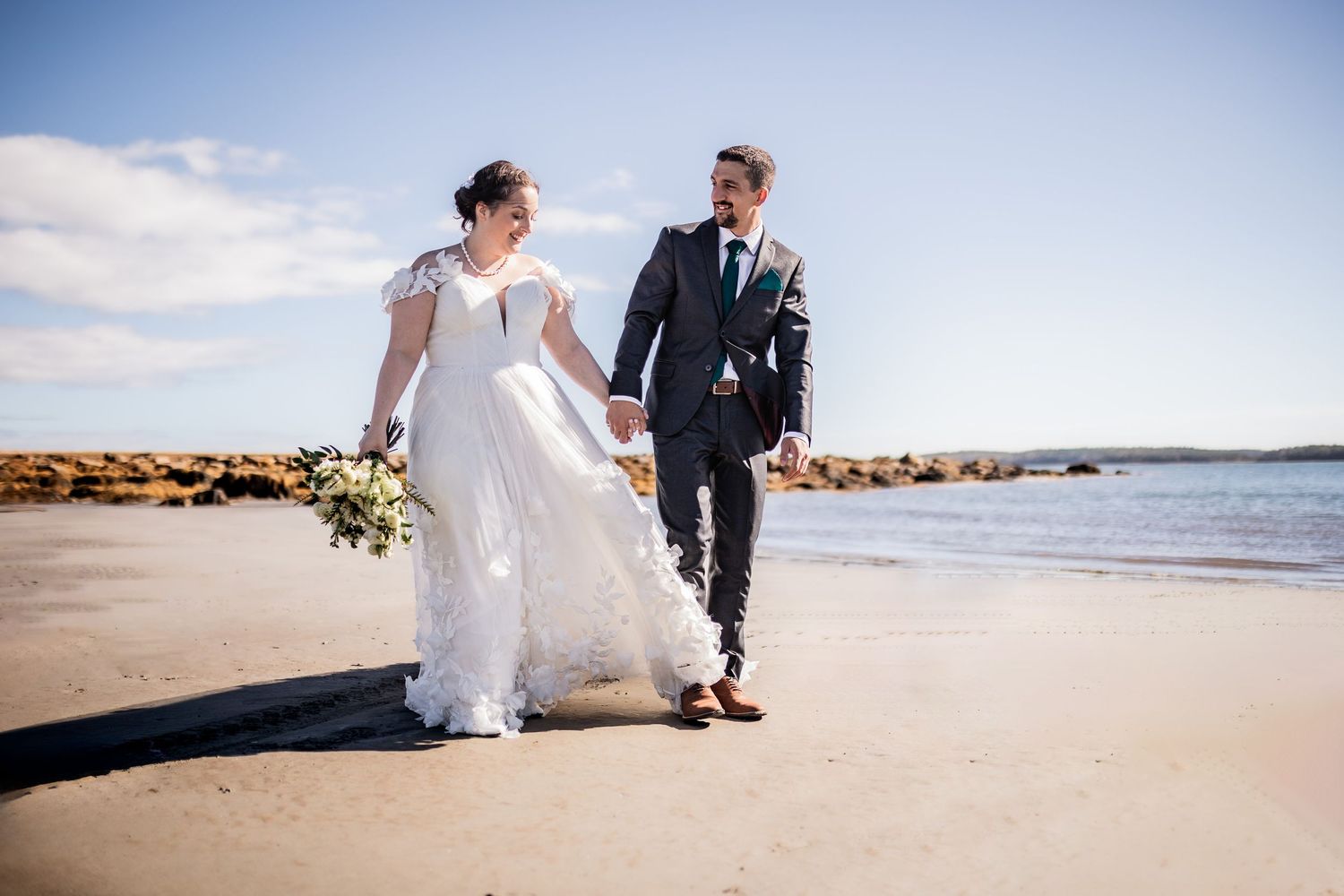 Wedding couple walking along beach shoreline on sunny day.