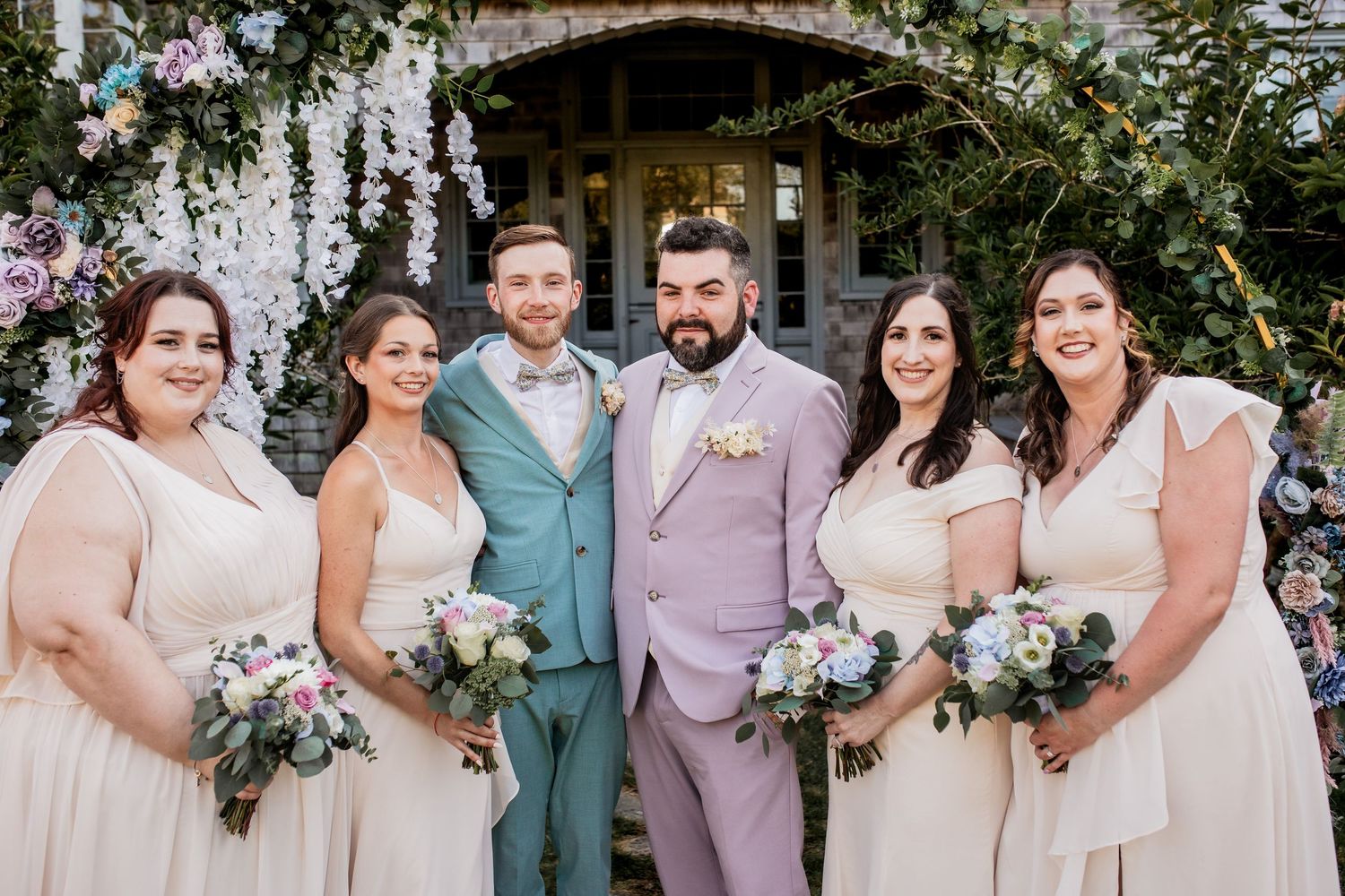 Wedding party in soft pastel attire poses together in front of floral-decorated entrance.