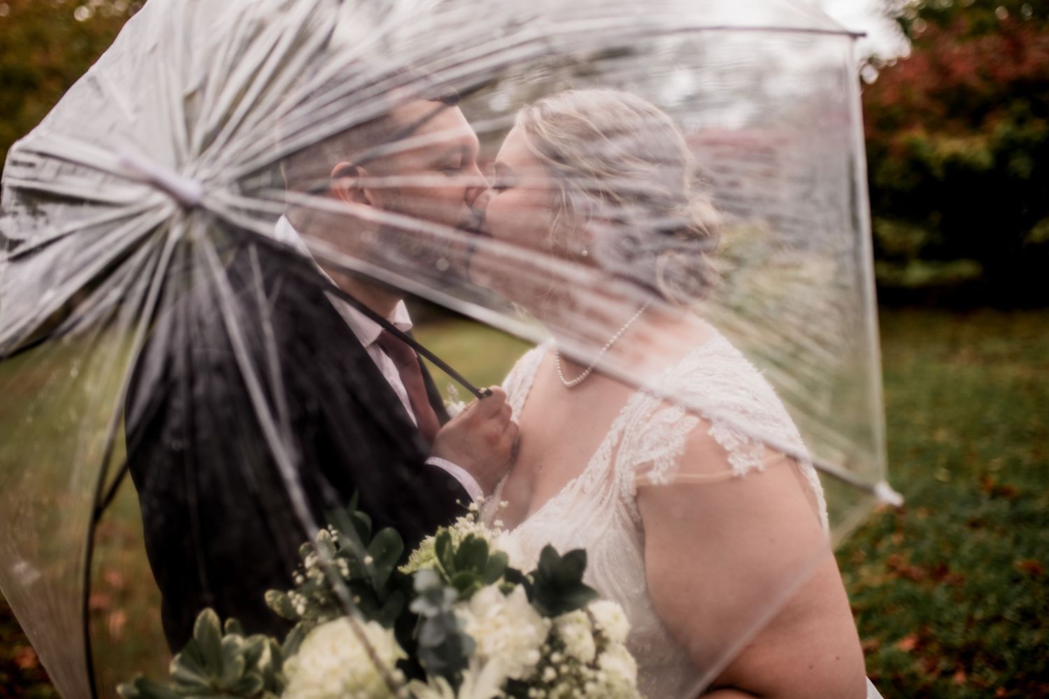Intimate moment captured under clear umbrella with white wedding bouquet.