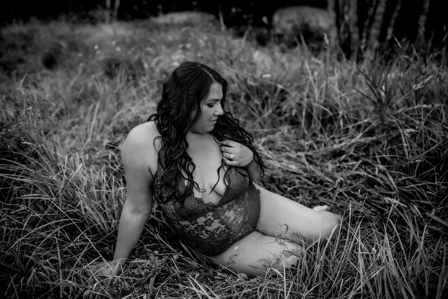 A black and white artistic boudoir portrait of someone sitting in tall grass wearing lace lingerie.