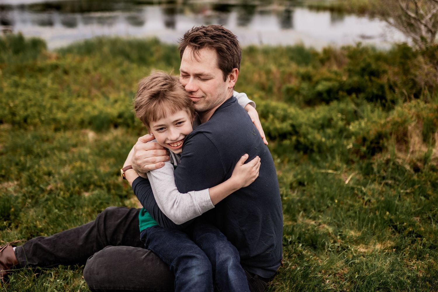 Candid outdoor portrait showing warm embrace between parent and child on grassy hillside.