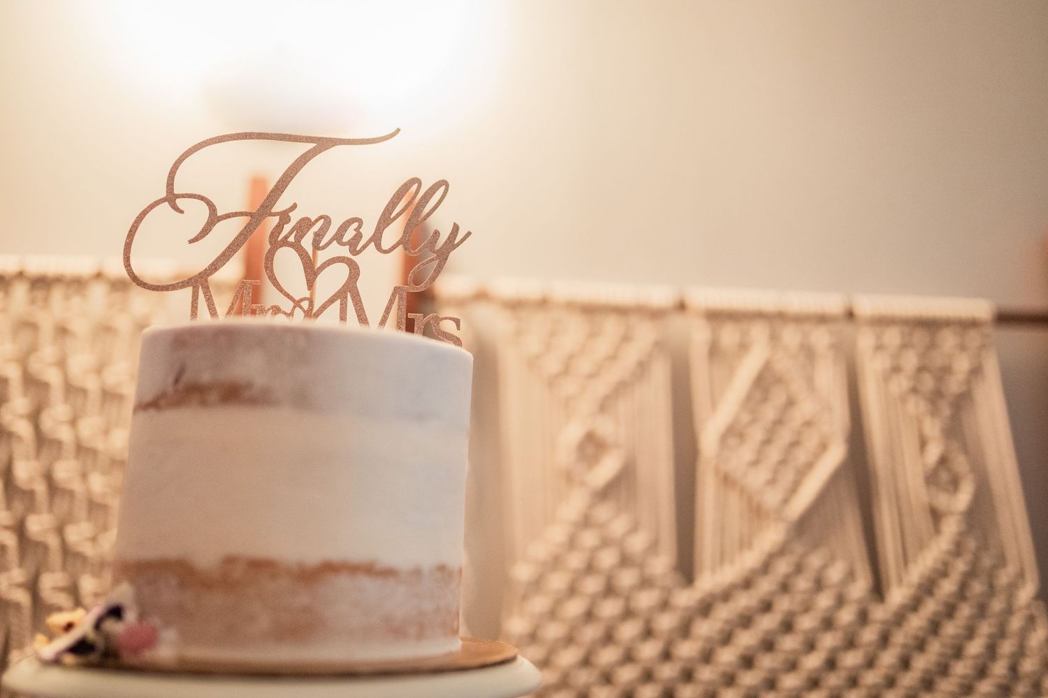 A minimalist semi-naked wedding cake with a 'Finally' rose gold topper and macrame backdrop.