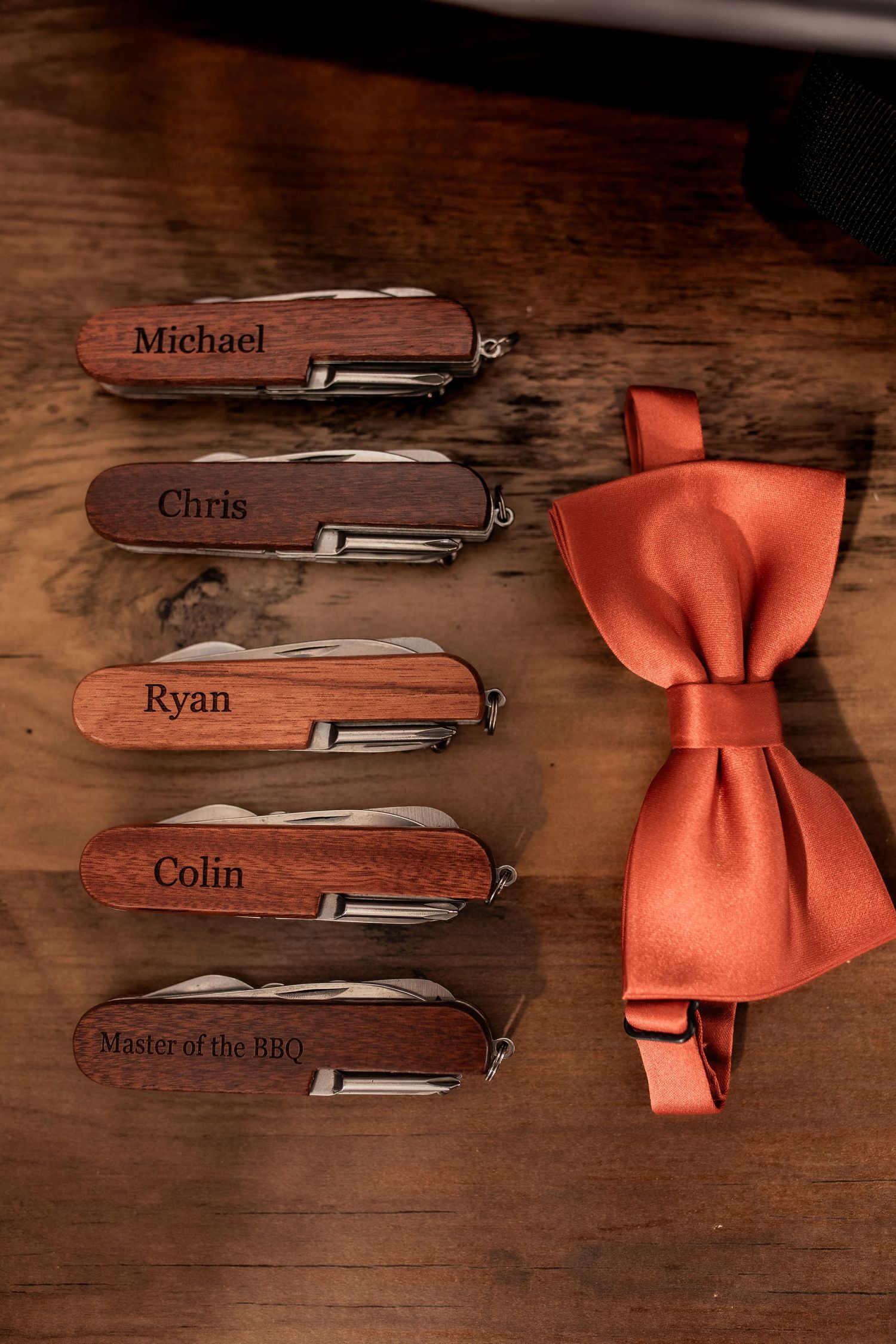 Personalized wooden groomsmen pocket knives and coral bow tie arranged on dark wooden surface.