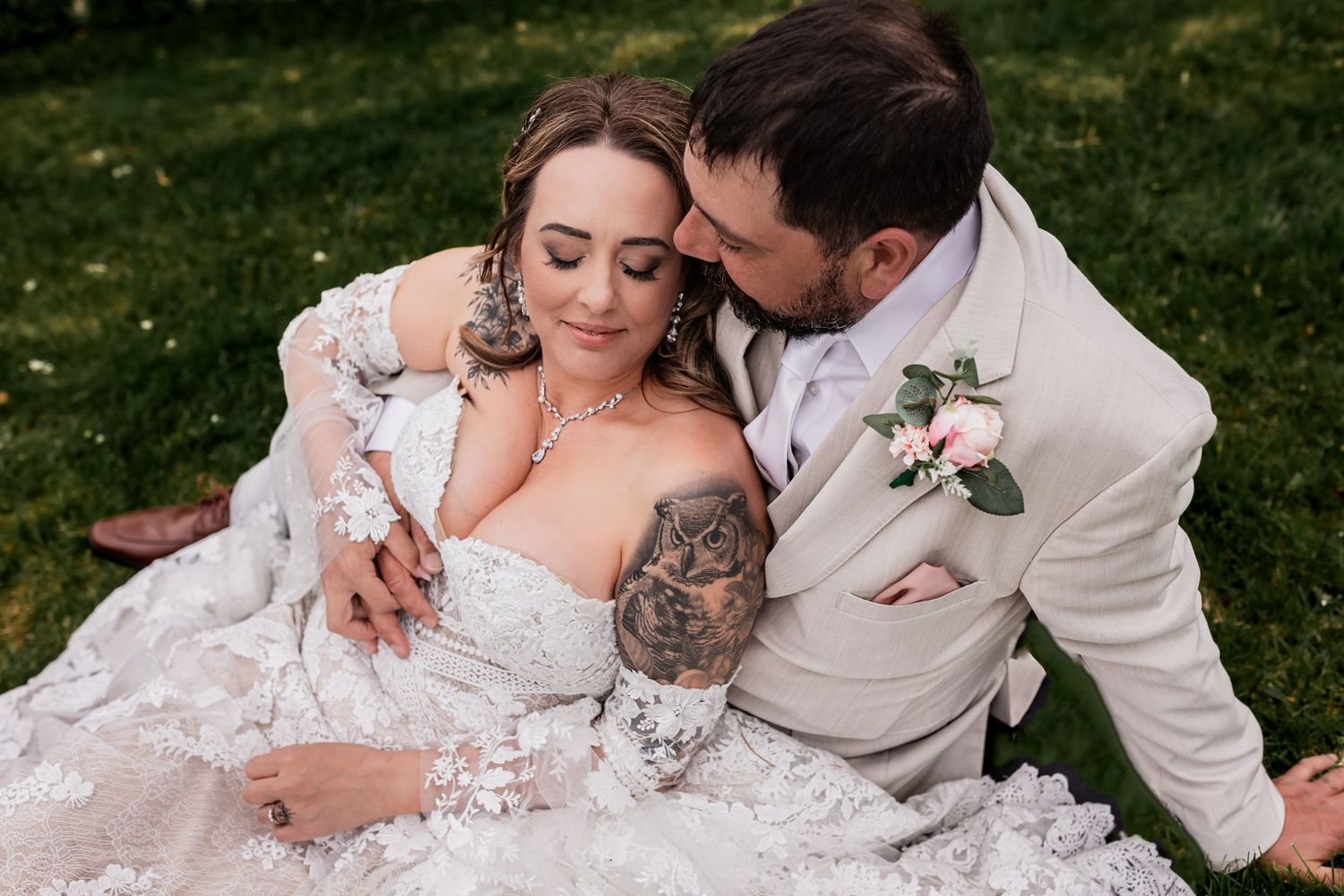 A romantic wedding couple embraces on grass, with the bride wearing a lace dress displaying her arm tattoo.