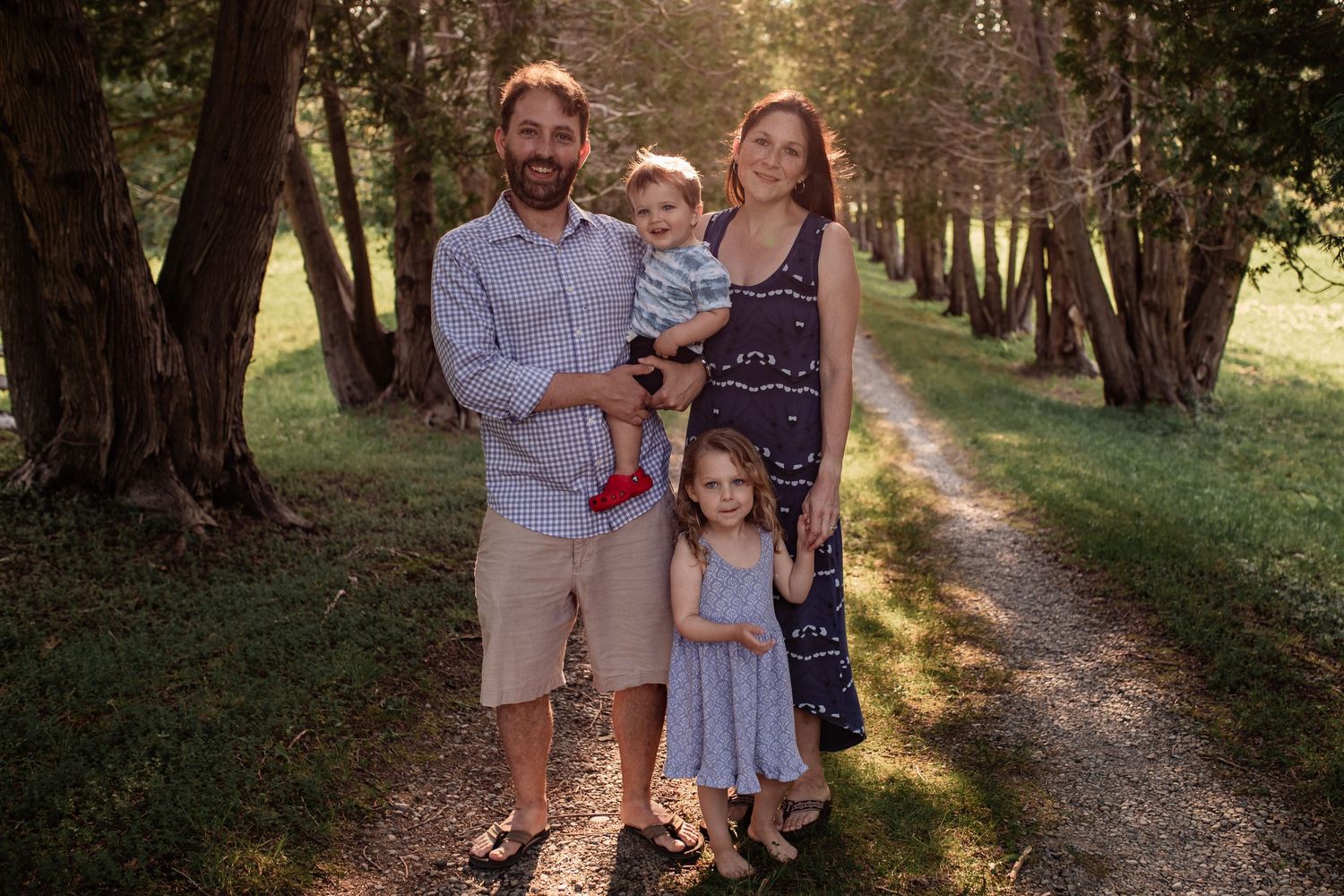 Family standing together on a tree-lined dirt path in warm evening sunlight.