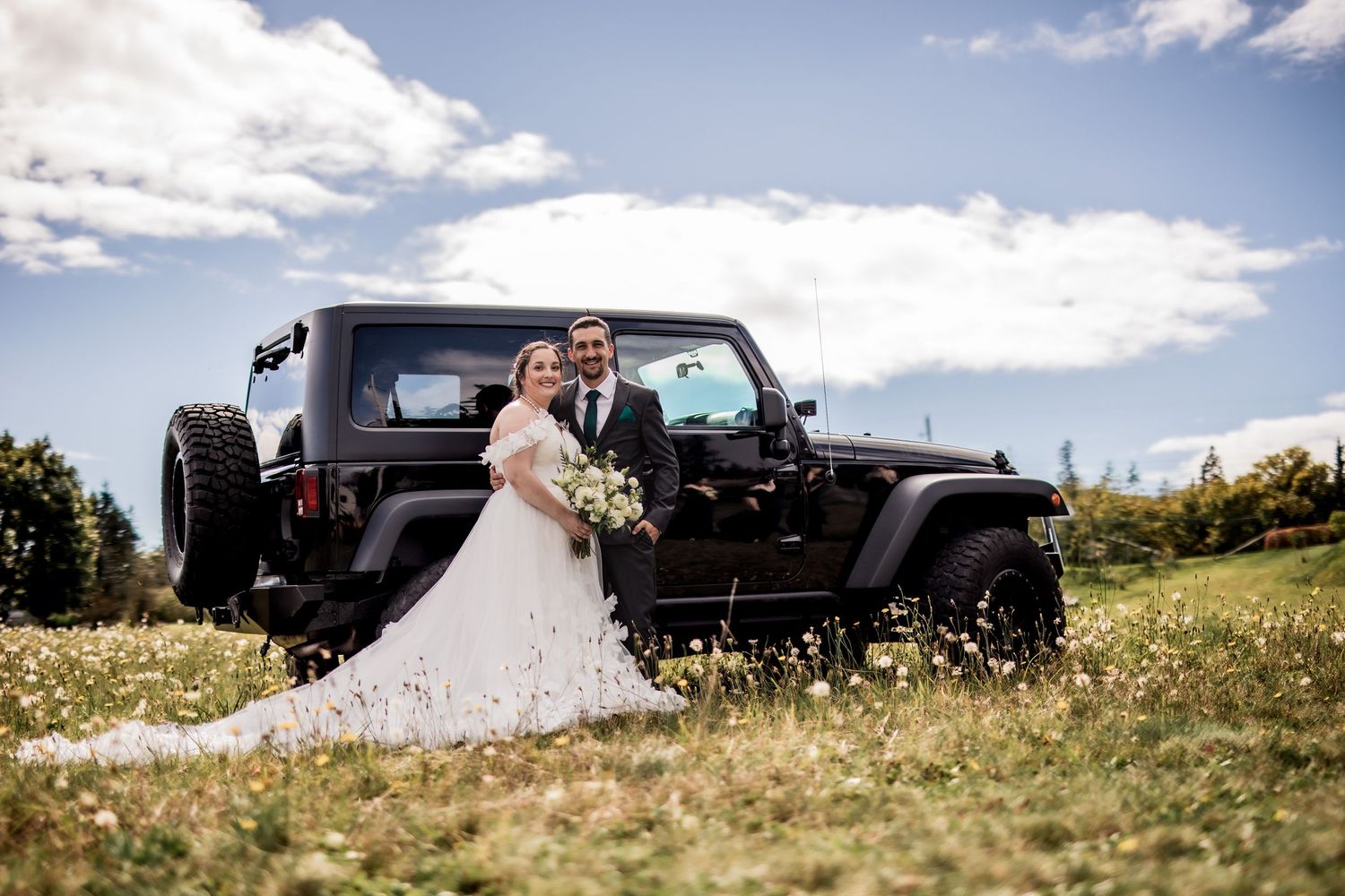 Wedding couple poses with black Jeep Wrangler in summer meadow.