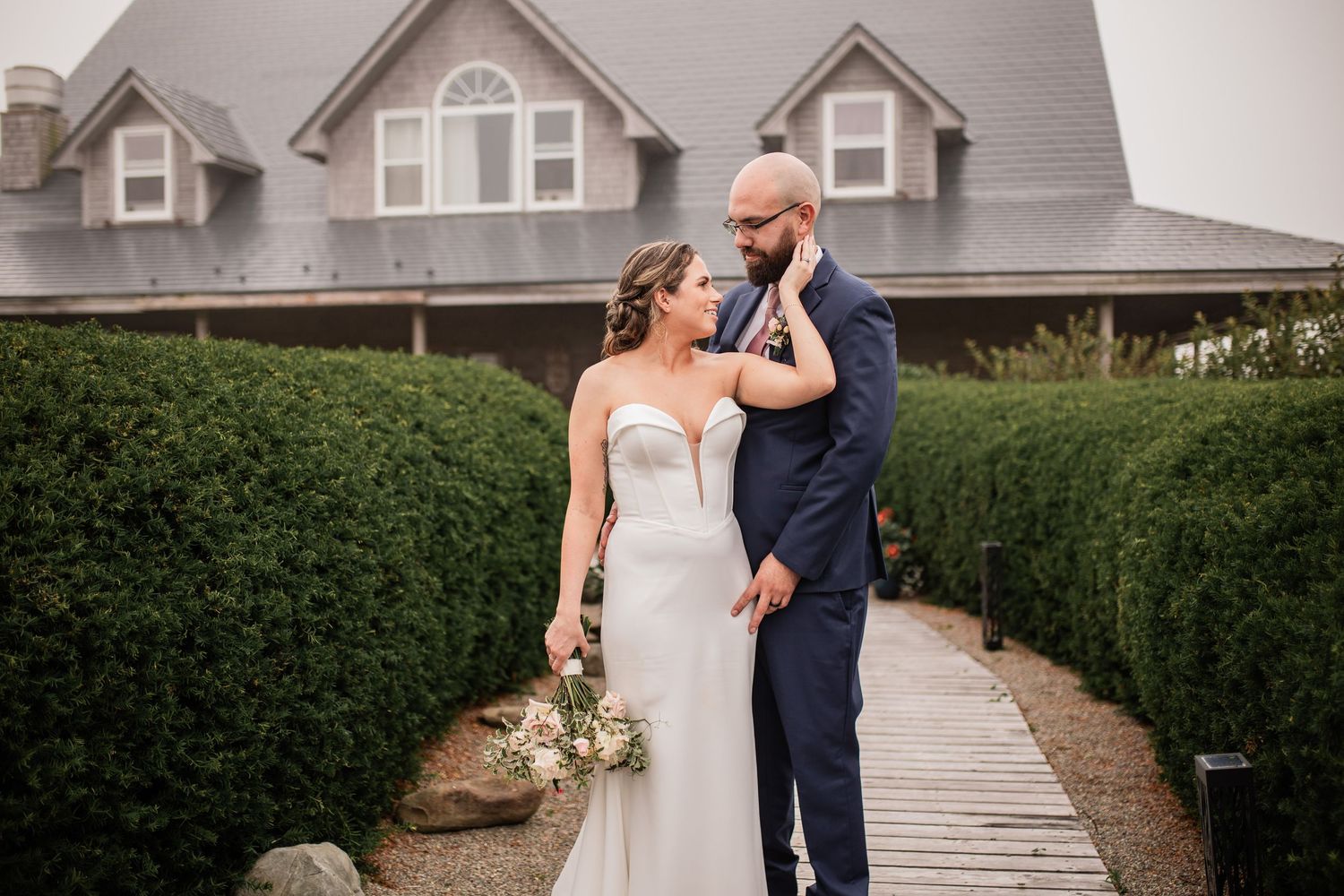 Newlyweds pose for portrait on walkway in front of Cape Cod style house with manicured hedges.