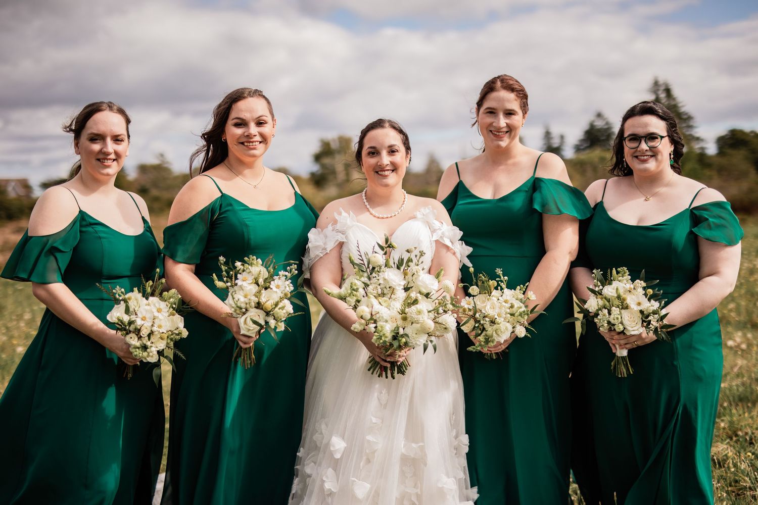 Bridal party in emerald green dresses poses with white bouquets outdoors.