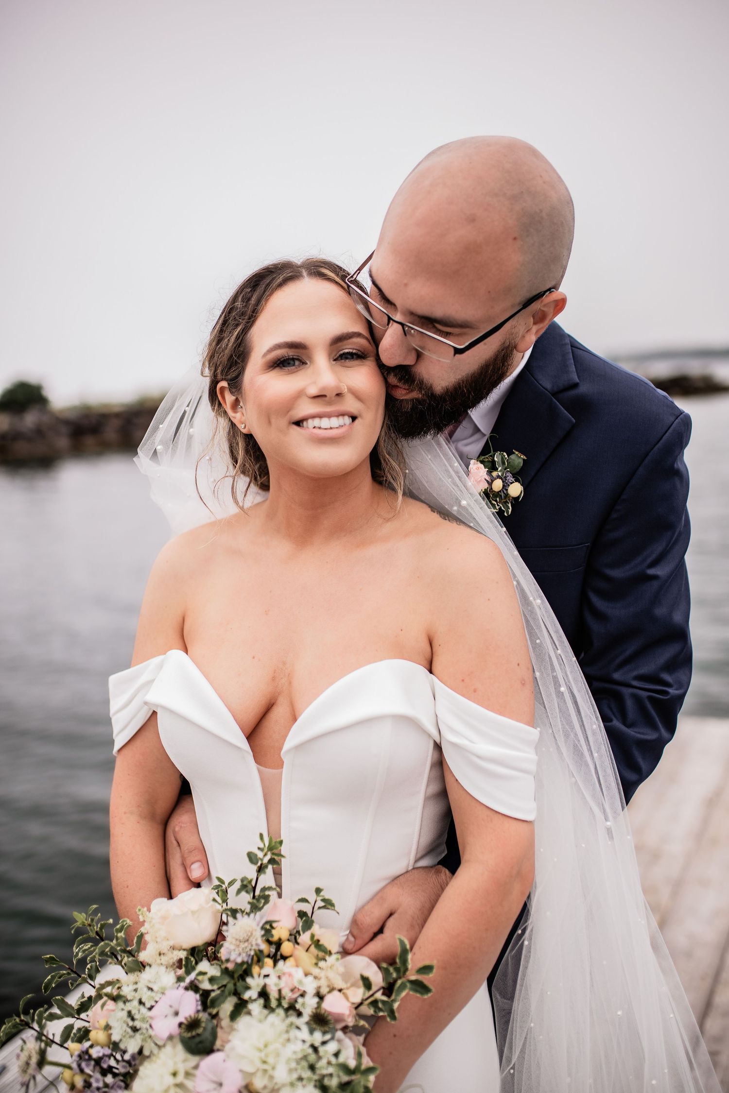 Tender moment between newlyweds by waterfront with flowing veil.