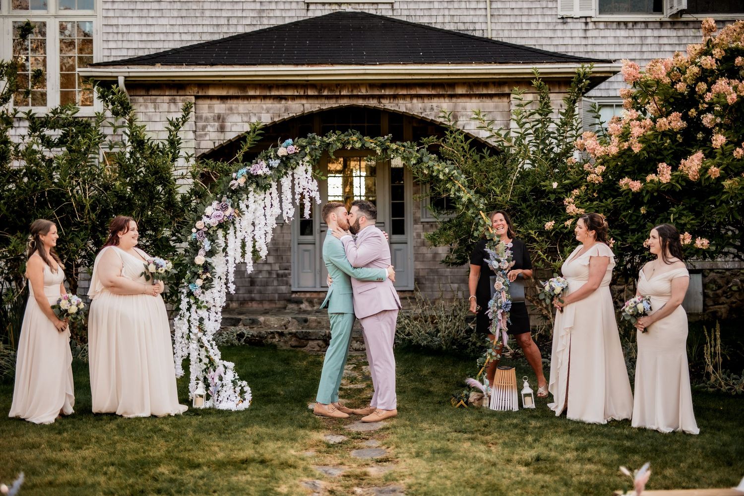 Wedding ceremony under floral arch with wedding party in pastel dresses and suits.