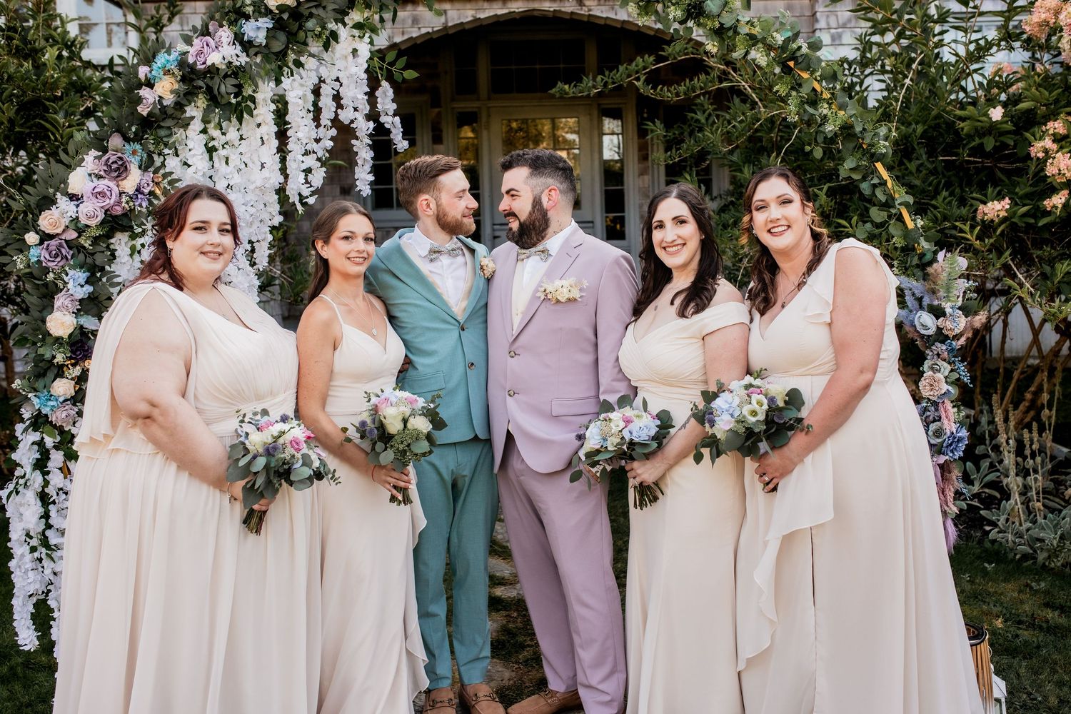 Wedding party wearing light colored dresses and suits stands together against garden backdrop.