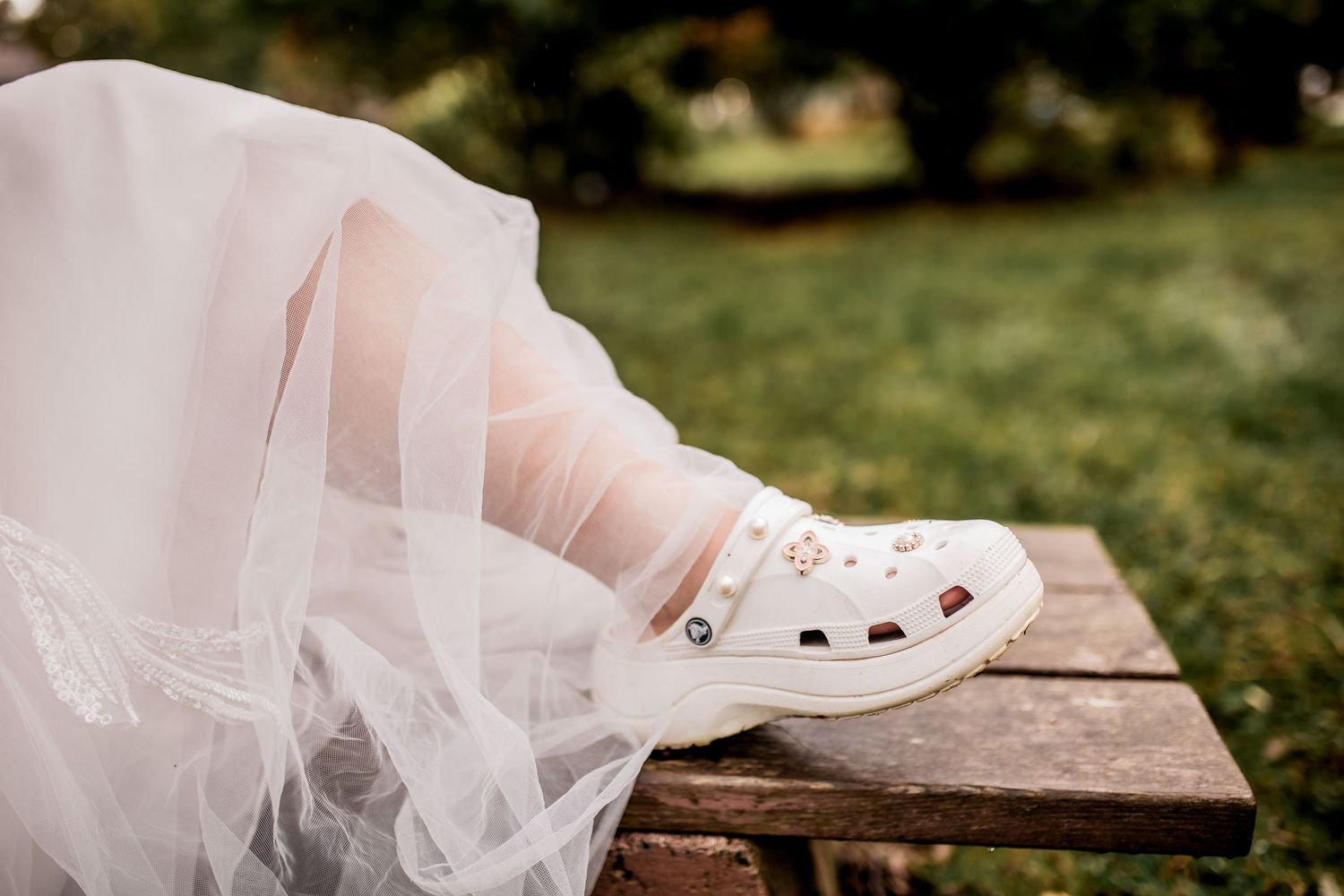 White wedding dress hem and Crocs shoes shown on rustic wooden bench outdoors.