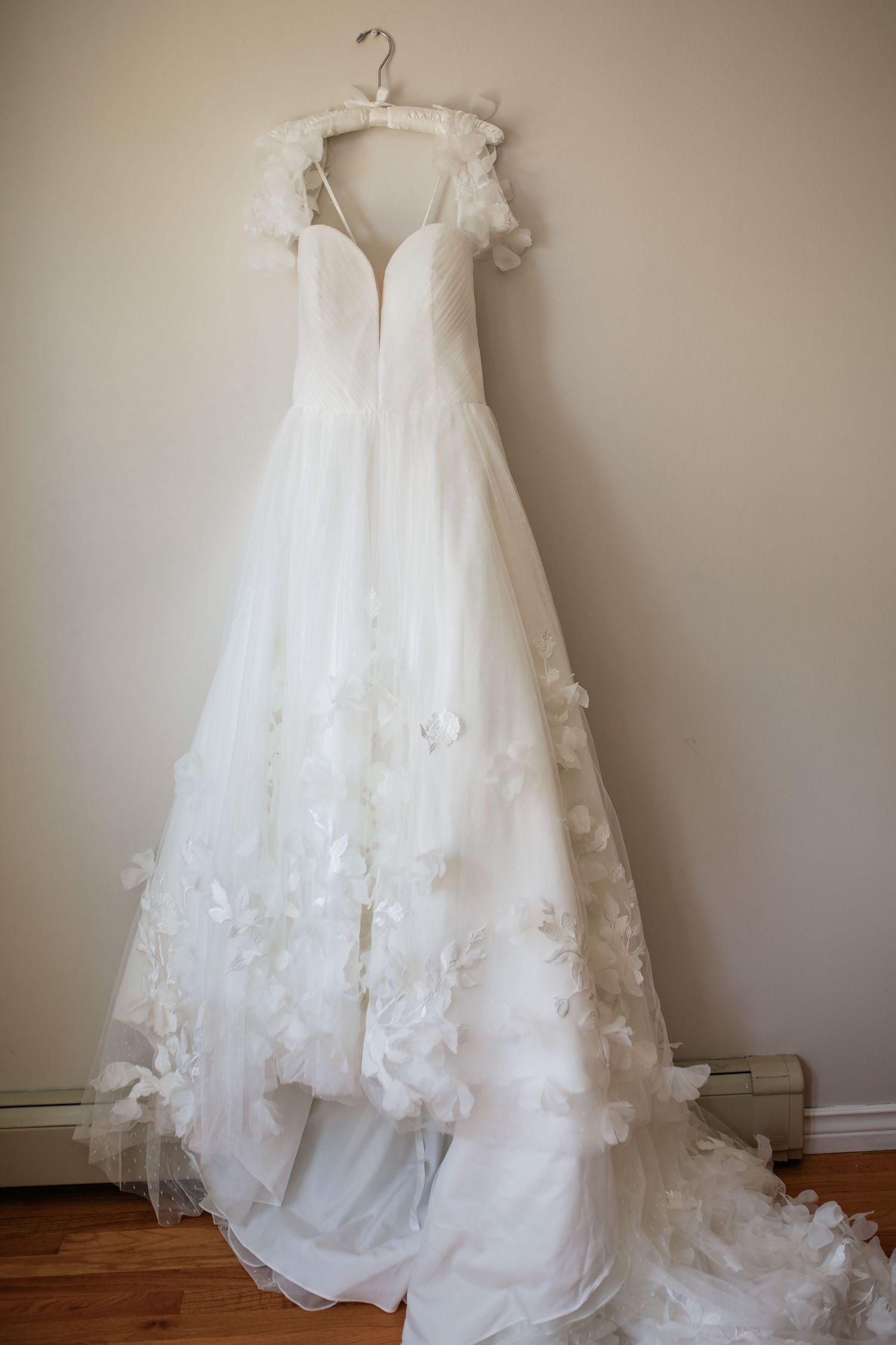 An elegant white wedding dress with lace details hanging on display against a neutral wall.