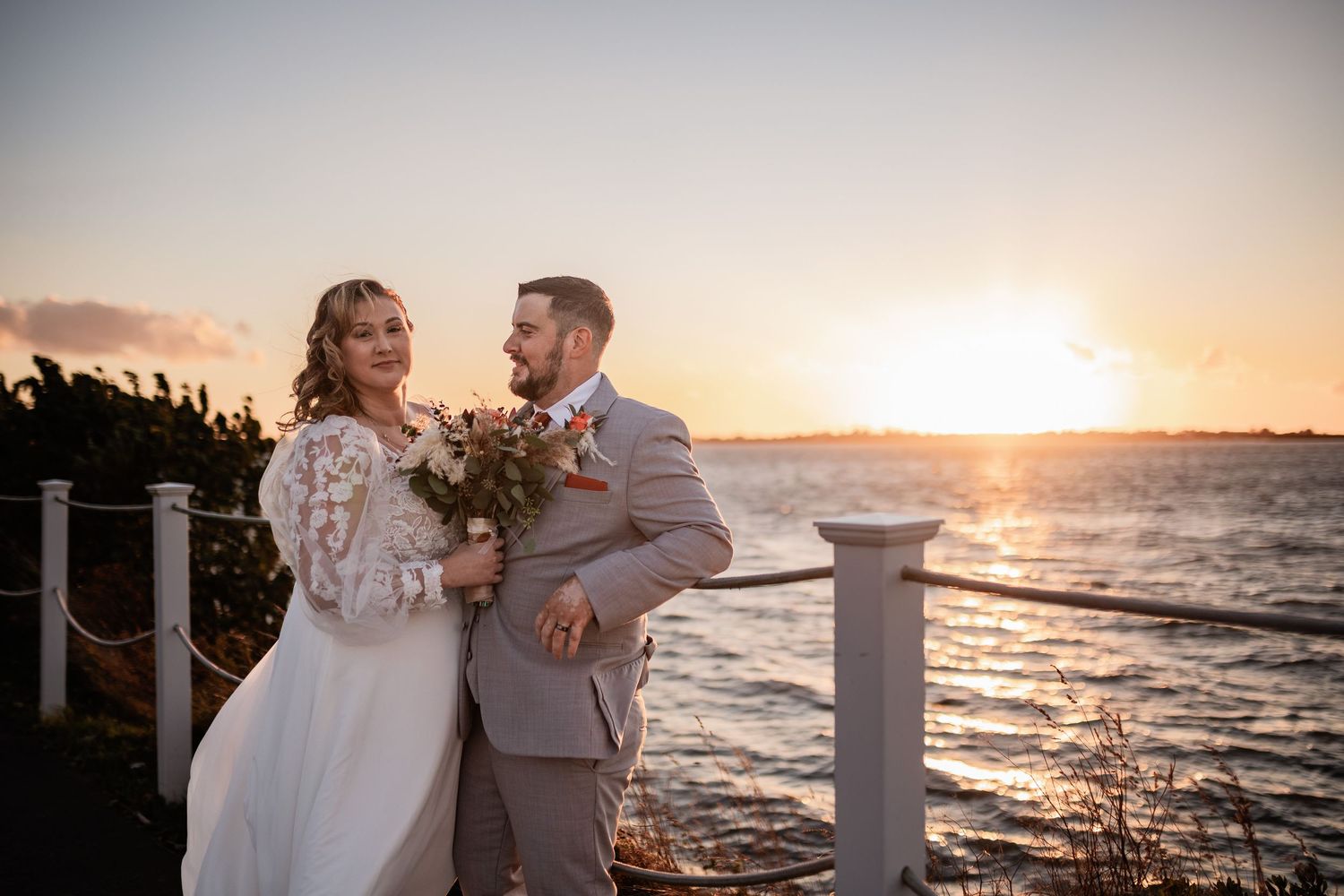 A romantic waterfront wedding scene with couple embracing by railing during golden sunset.