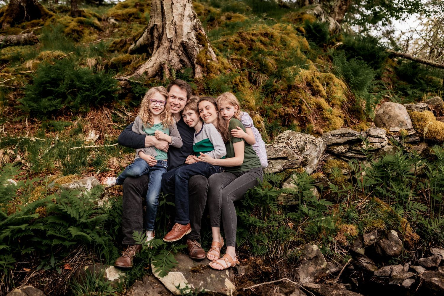 Group sitting together on natural rock formation in wooded area during outdoor photo session.