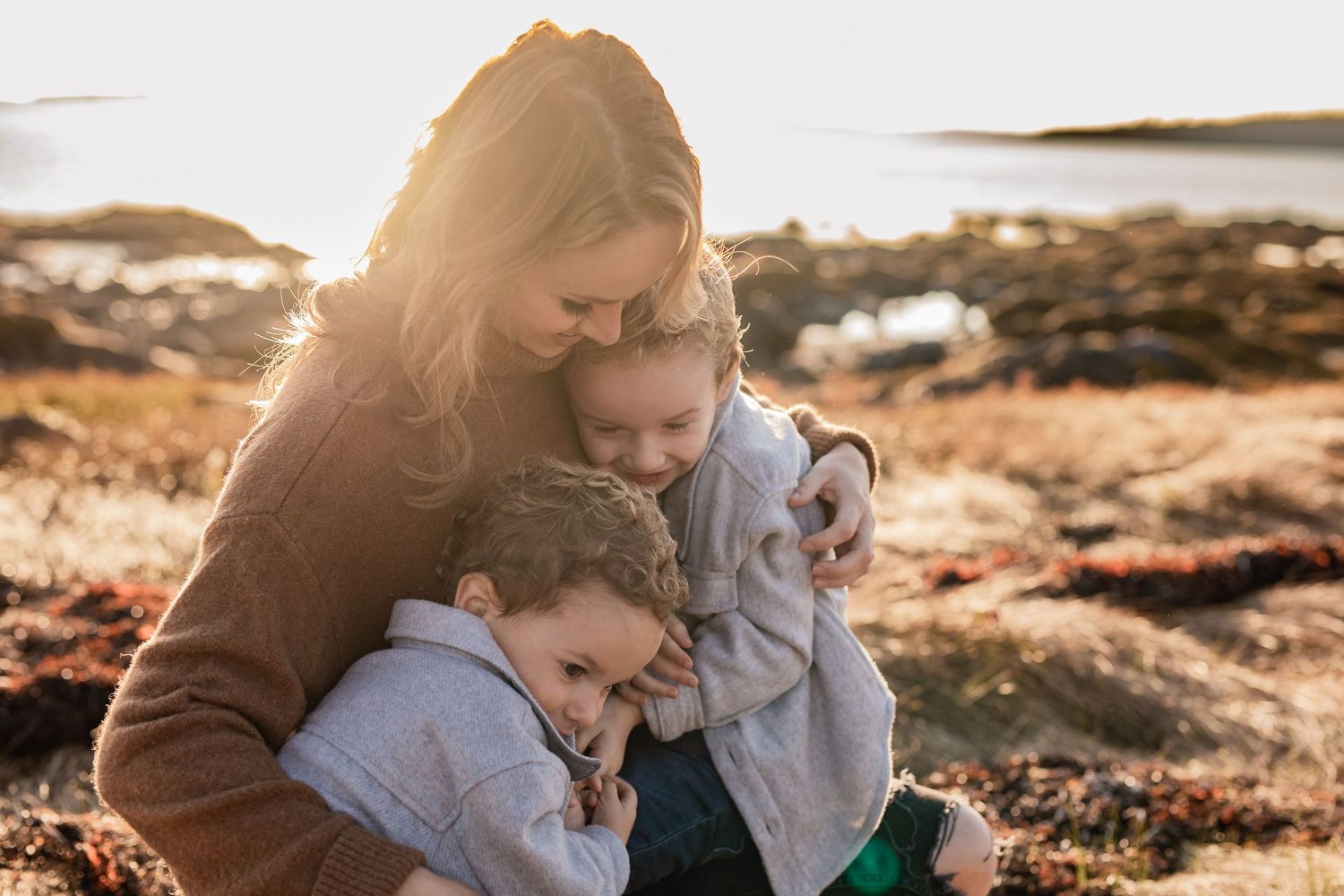 A warm sunset moment as someone embraces two others on a rocky coastal shoreline.