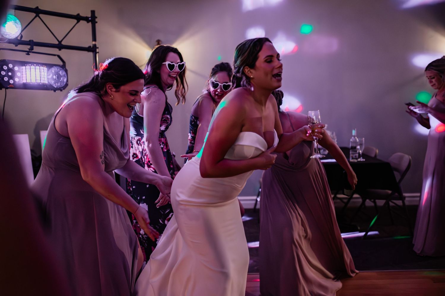 Wedding party dancing energetically at reception under purple mood lighting.