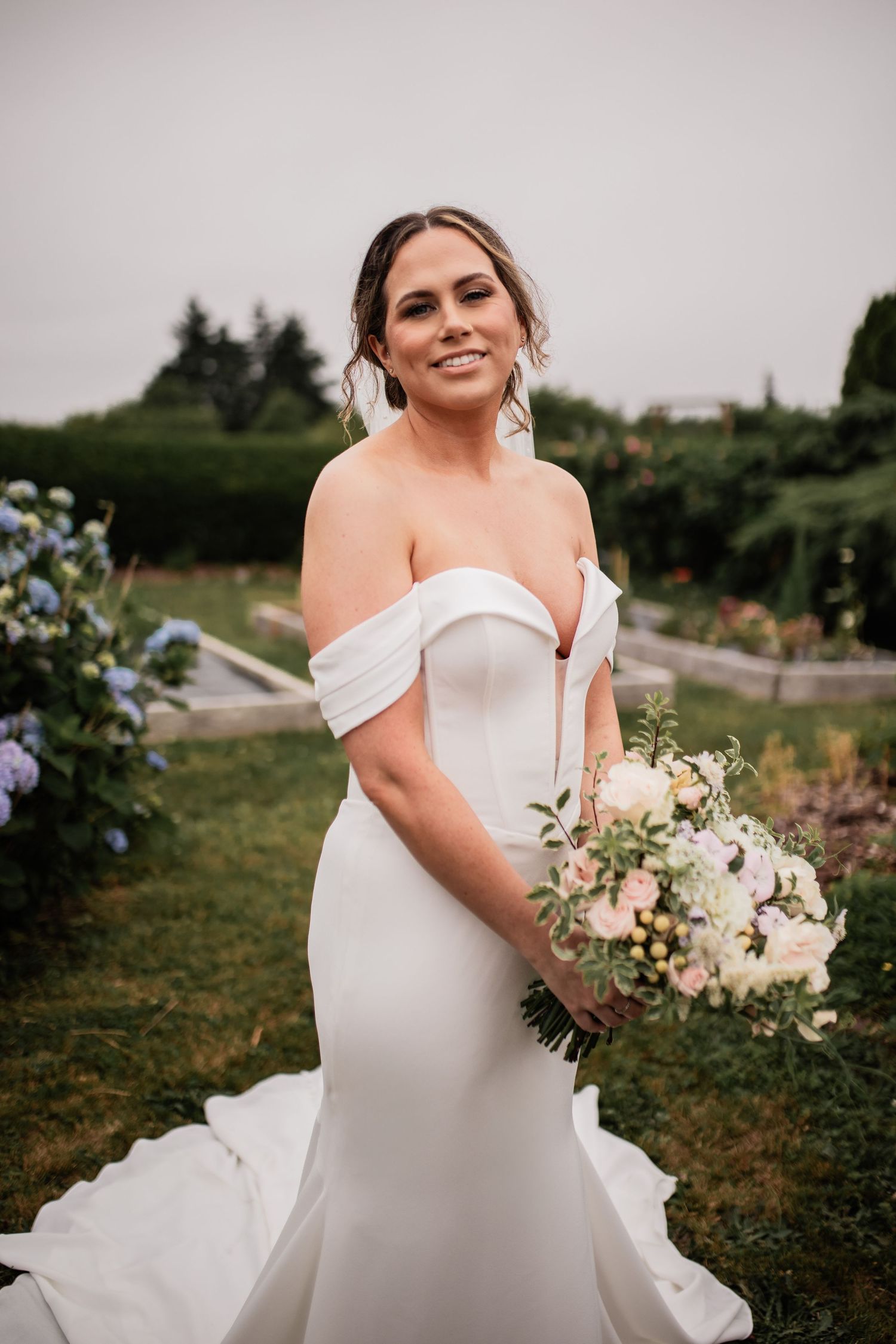 Elegant off-shoulder wedding dress shown in outdoor garden setting with bouquet.