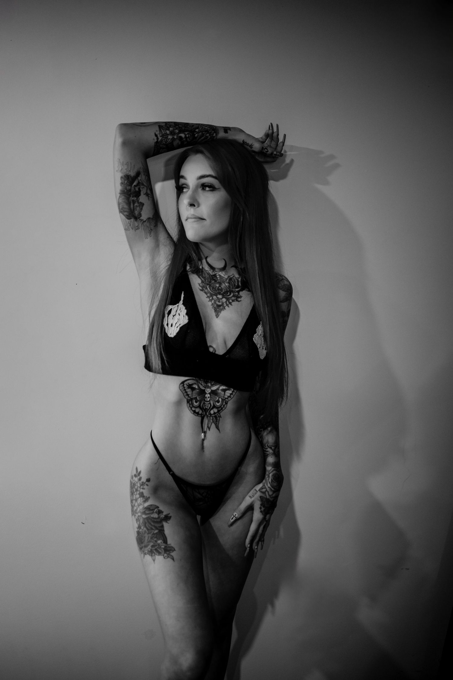 Black and white artistic portrait showcasing intricate tattoo art and black lingerie against plain wall.