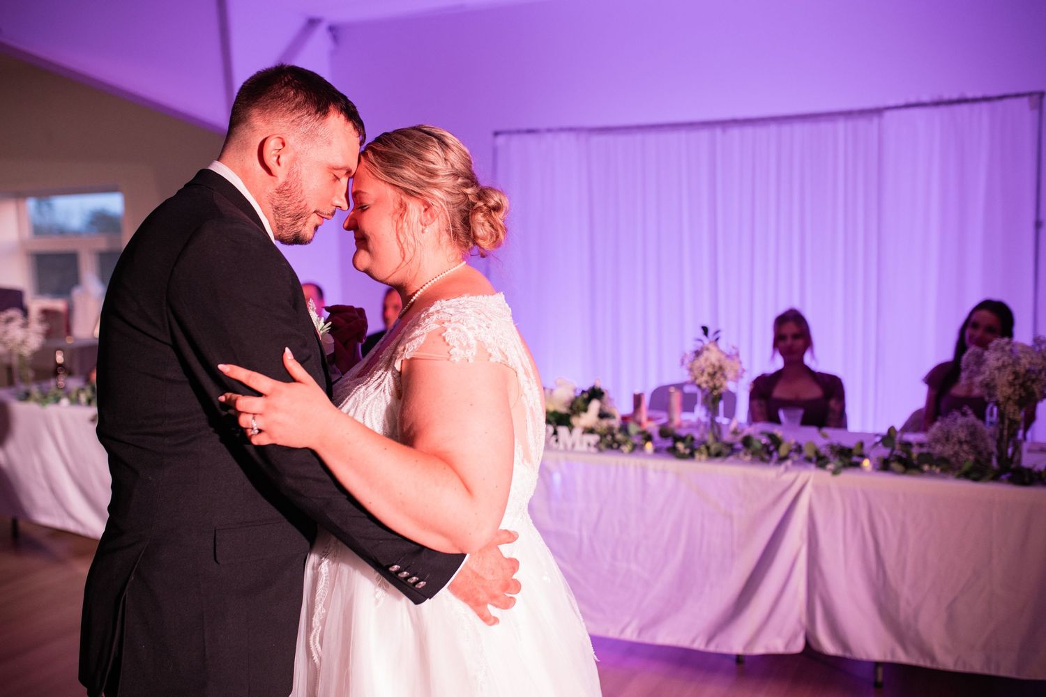 Couple shares romantic dance under purple lighting at wedding reception.
