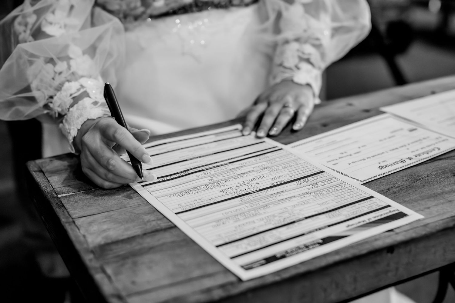 Close up of hands signing marriage license documents on wooden table.